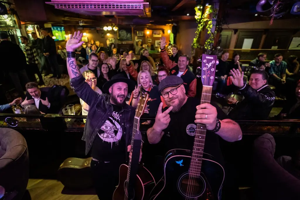 Ryhmäkuva tunnelmallisesta pubista. Etualalla kaksi  kitaristia ja yleisöä taustalla.