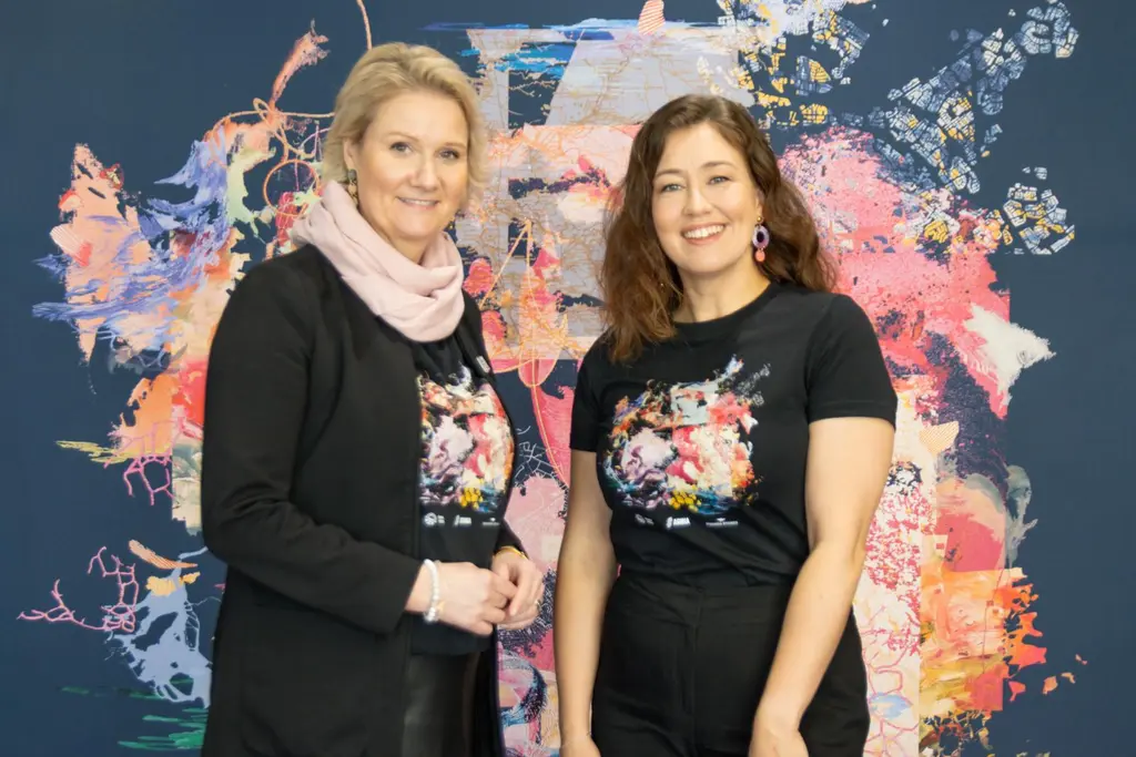 Kaksi naista katsoo hymyillen kameraan. He seisovat suuren Mielestäni-kuosin edessä.