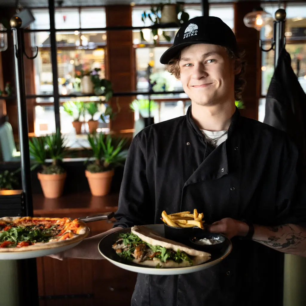 Nuori ravintolakokki hymyilee kaksi ruoka-annosta käsissään.