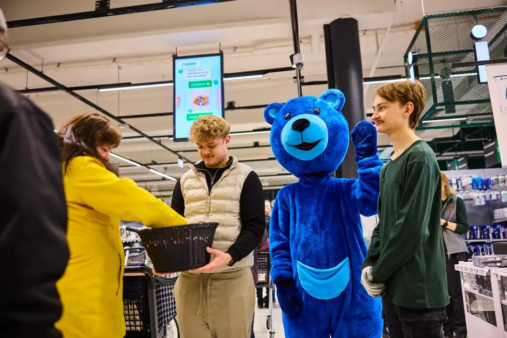 Kaksi henkilöä jakamassa asiakkaille karkkeja ja kameralle vilkuttava Sini-nalle.