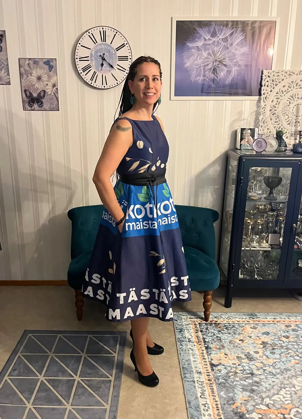 Nainen seisoo kasvot kameraan. Hänellä on päällään mekko, jonka kuosi on S-ryhmän Kotimaista-tuotesarjasta.