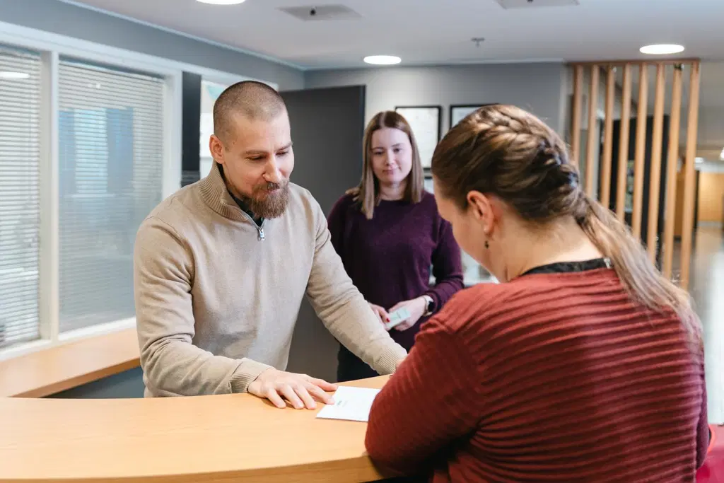 Kaksi konttorin työntekijää seisoo infon tiskin takana ja neuvoo henkilöä lomakkeen täytössä. 