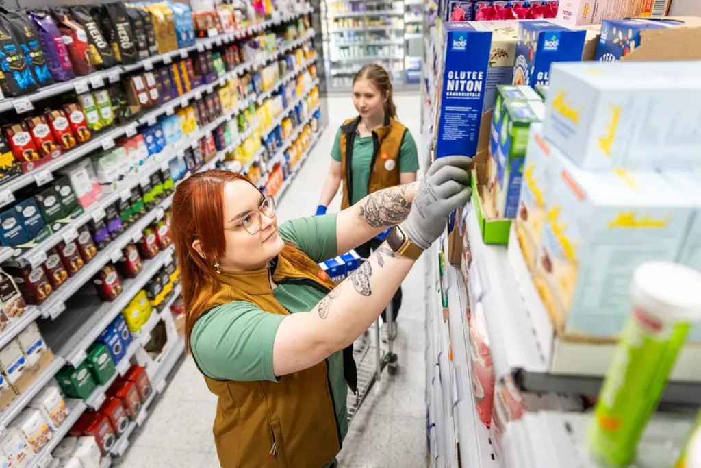 Ruokakaupan myyjä hyllyttää tuotteita. Työkaveri seisoo taustalla, edessään hänellä on kärryt, joissa on tuotteita.