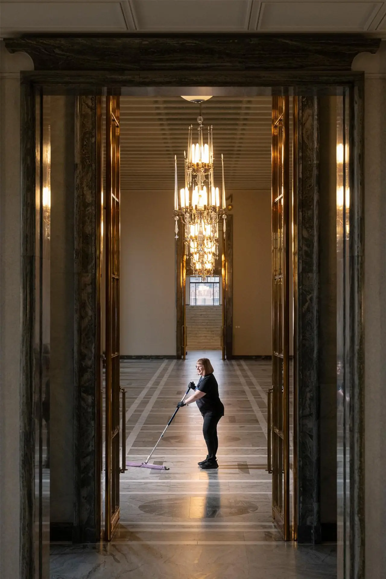 Laitoshuoltaja Minna Sepponen siivoaa eduskuntatalon lattiaa huoneessa, jossa on suuri kristallikruunu.