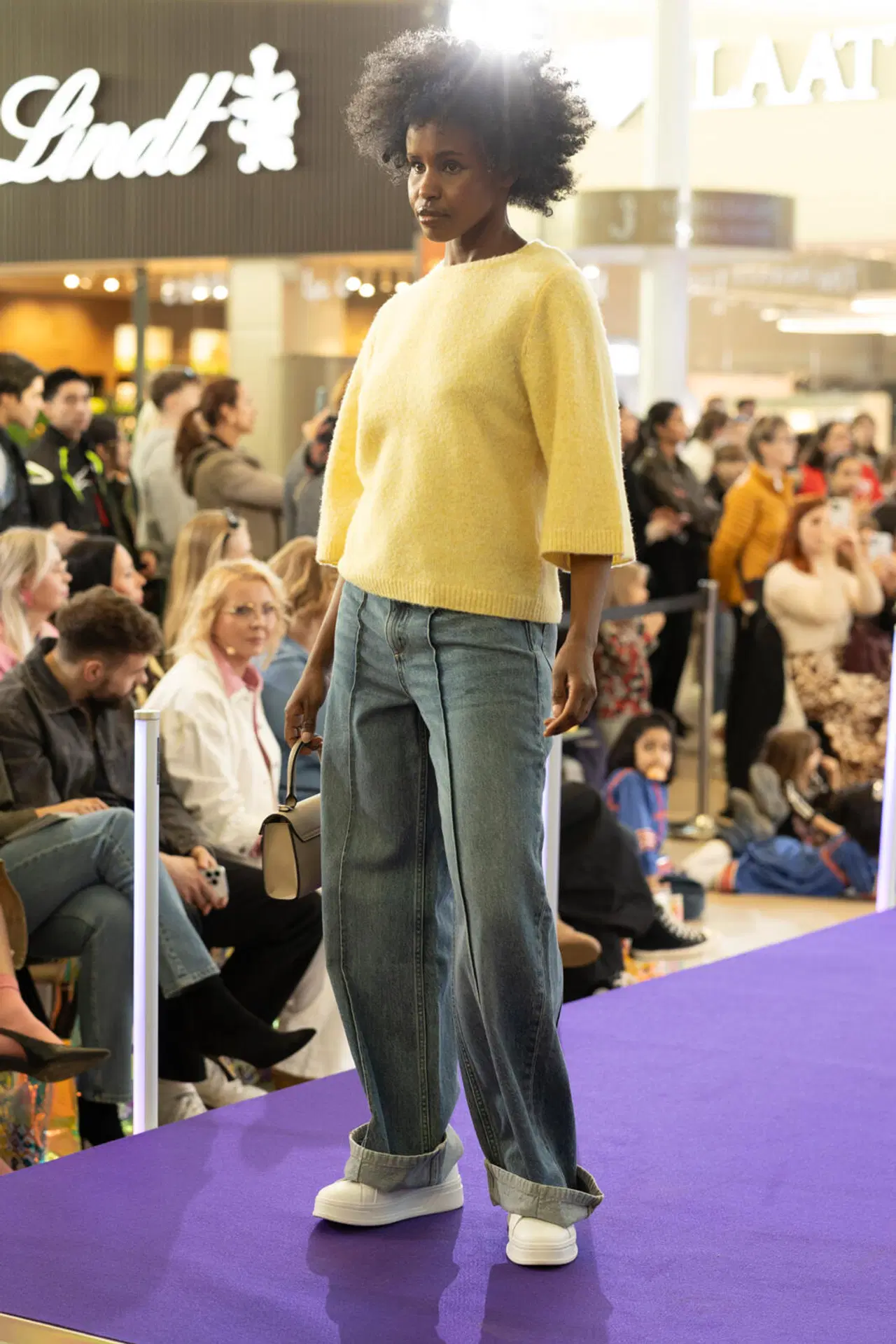 Malli kävelee catwalkilla keltaisessa neuleessa, leveissä farkuissa ja valkoisissa tennareissa, pientä laukkua kantaa.