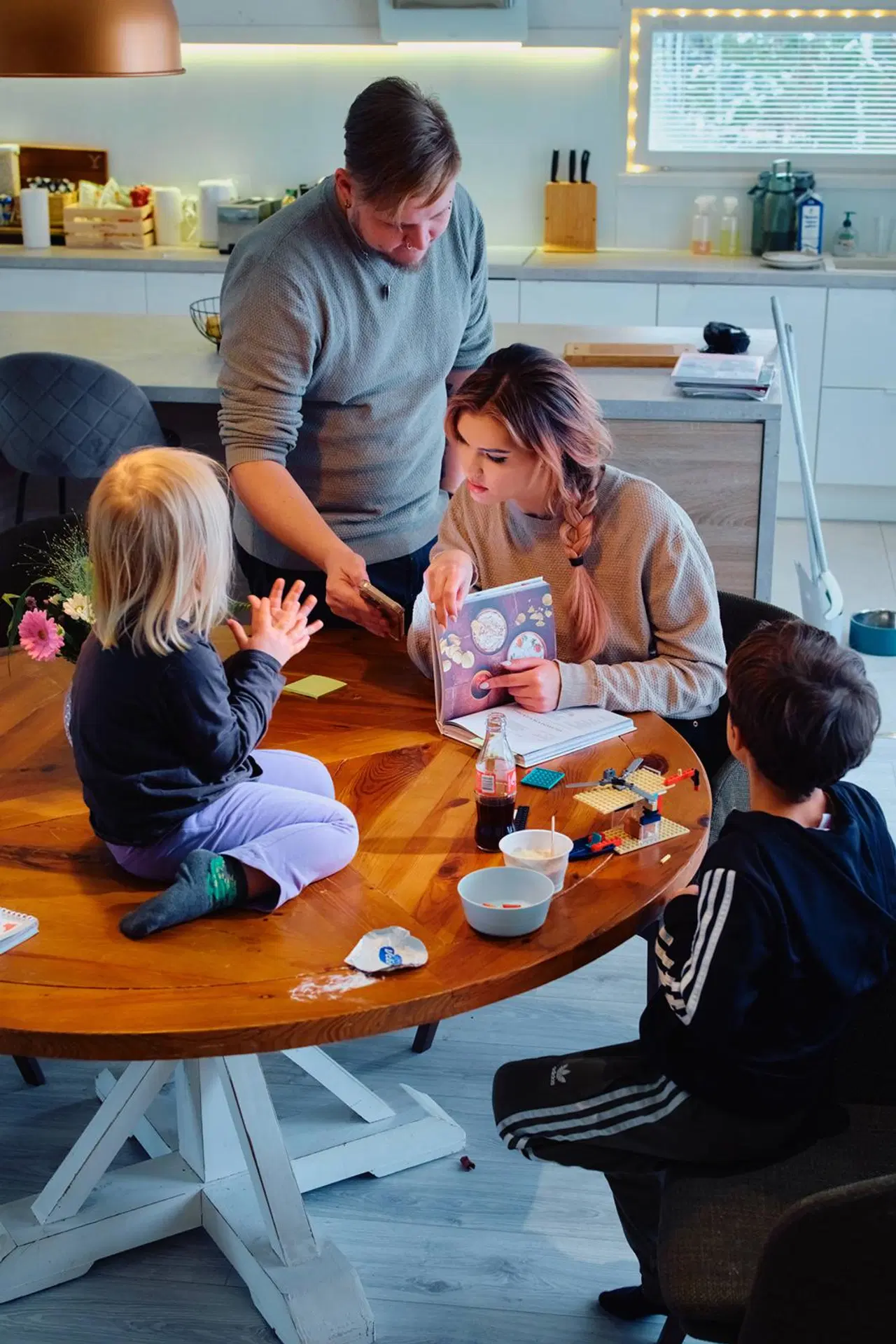 Kangaskortetin perhe pyöreän pöydän ääressä: Tommi näyttää Marille puhelimensa ruutua, Marialla kädessä kirja, lapsi istuu pöydän päällä ja toinen tuolilla.