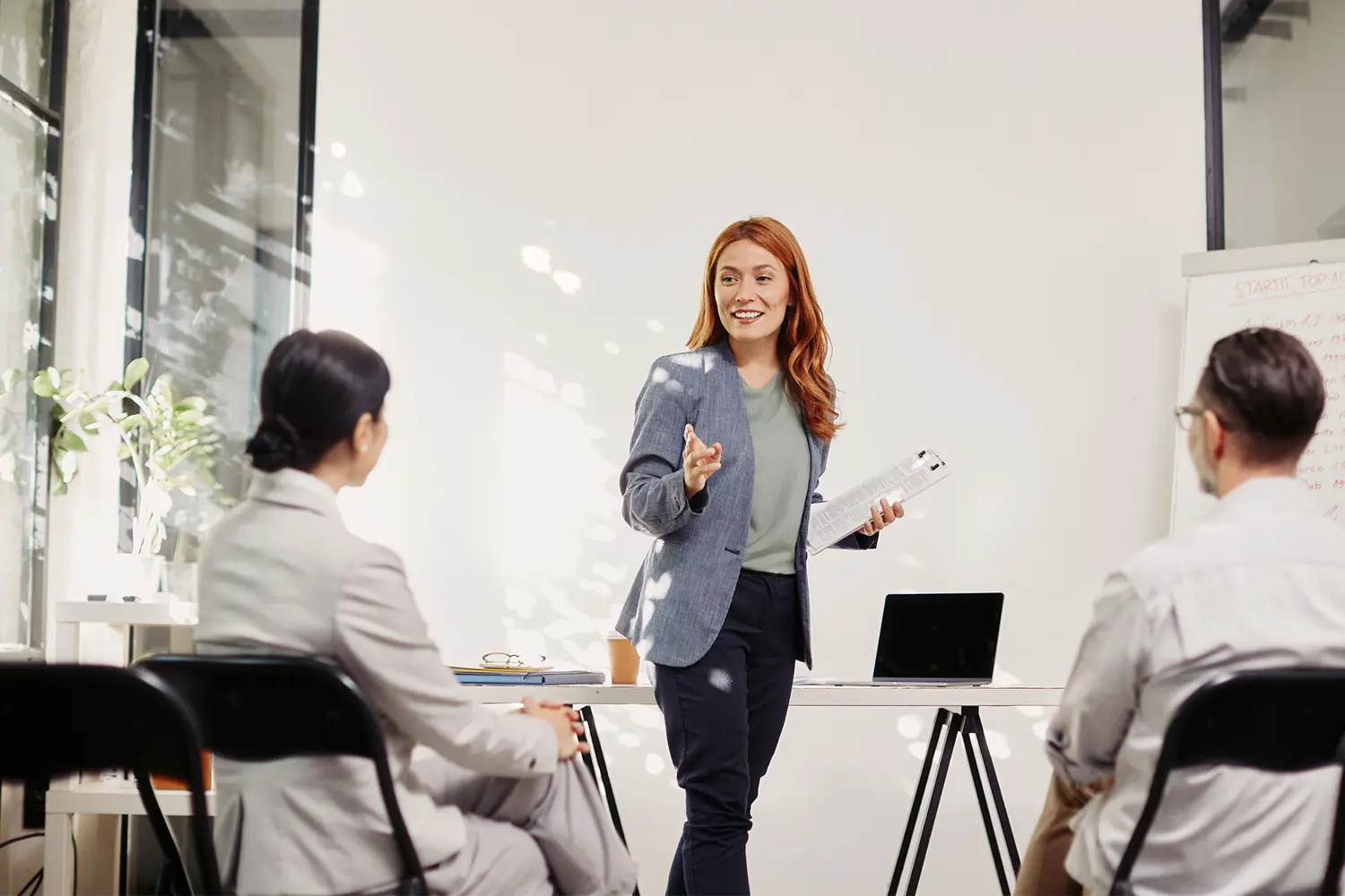 Nainen seisoo hymyillen työpöydän edessä ja puhuu kahdelle istuvalle ihmiselle.