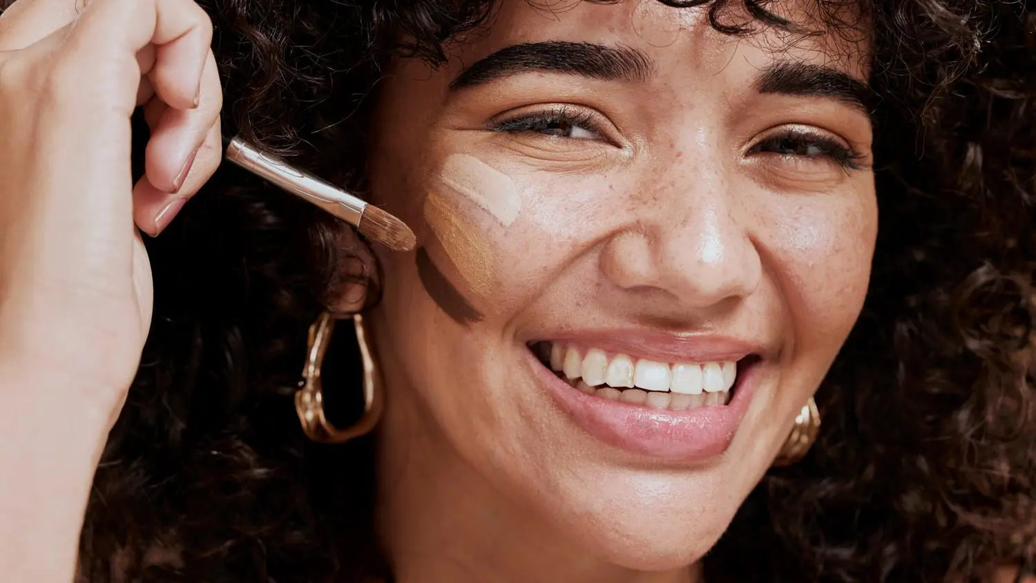 Hymyilevä nainen, jonka poskessa on raidat kolmea eri sävyistä meikkivoidetta. Naisella on meikkisivellin kädessään.
