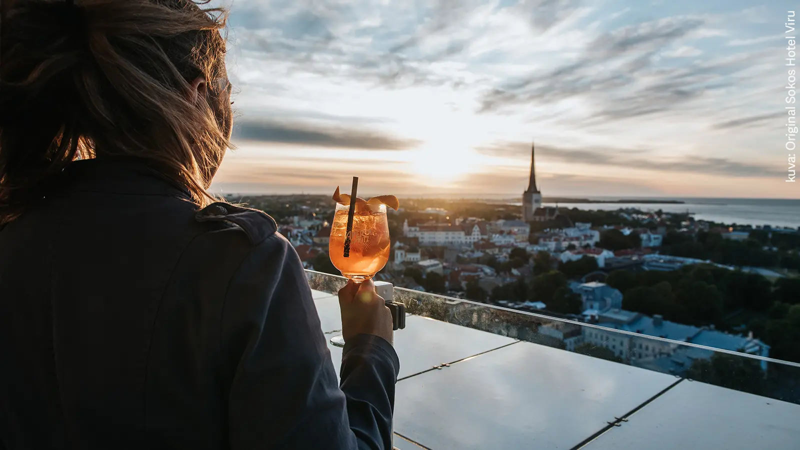 Nainen seisoo selin kameraan kattoterassilla drinkki kädessä. Taustalla Tallinnan kaupunkimaisema.