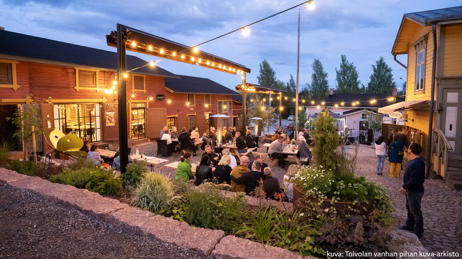 Toivolan vanhan pihan puutaloja ja iltaa viettäviä ihmisiä tunnelmallisessa valaistuksessa.