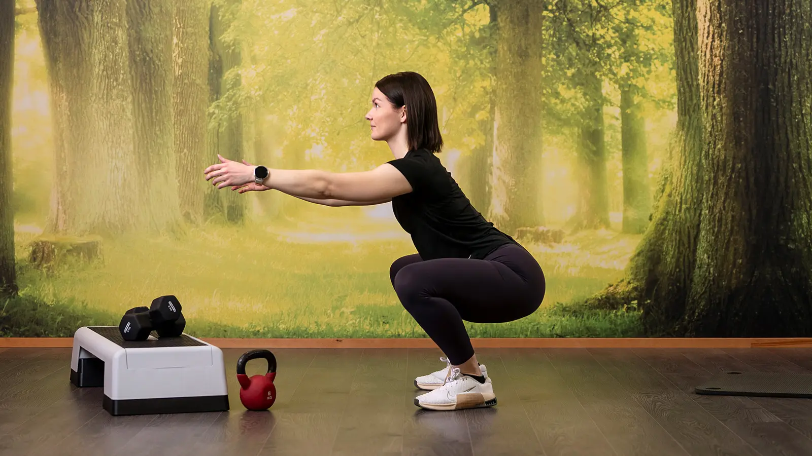 Nainen kyykkää, edessä steppilauta, käsipainot ja kahvakuula.