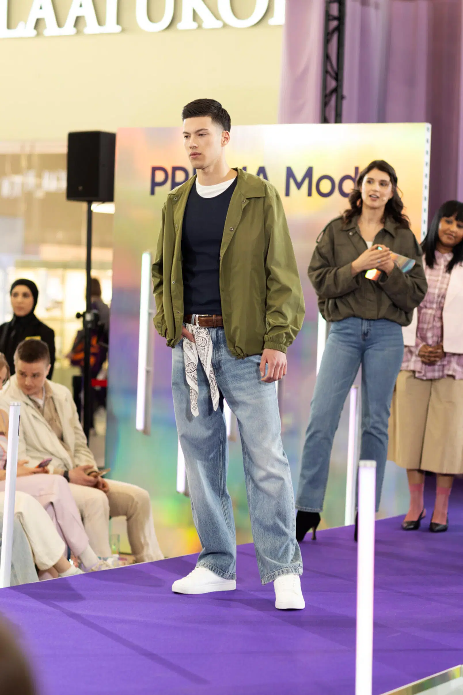 Mies mallikävelyllä: vihreä takki, tummansininen paita, vaaleat farkut ja valkoiset lenkkarit violetilla catwalkilla.