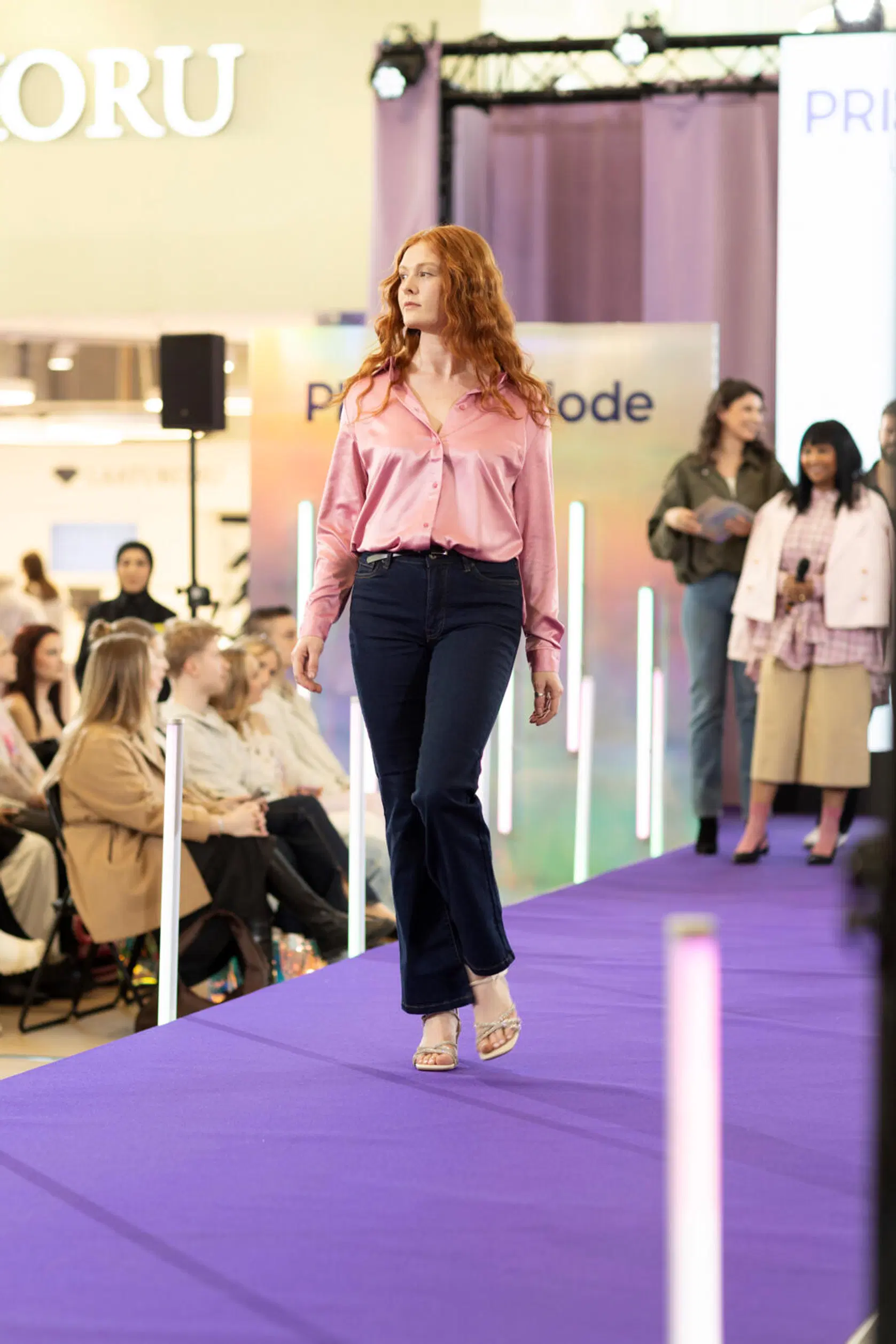 Malli kävelee catwalkilla vaaleanpunaisessa silkkipaidassa ja tummissa farkuissa, violetti lava, yleisö taustalla.