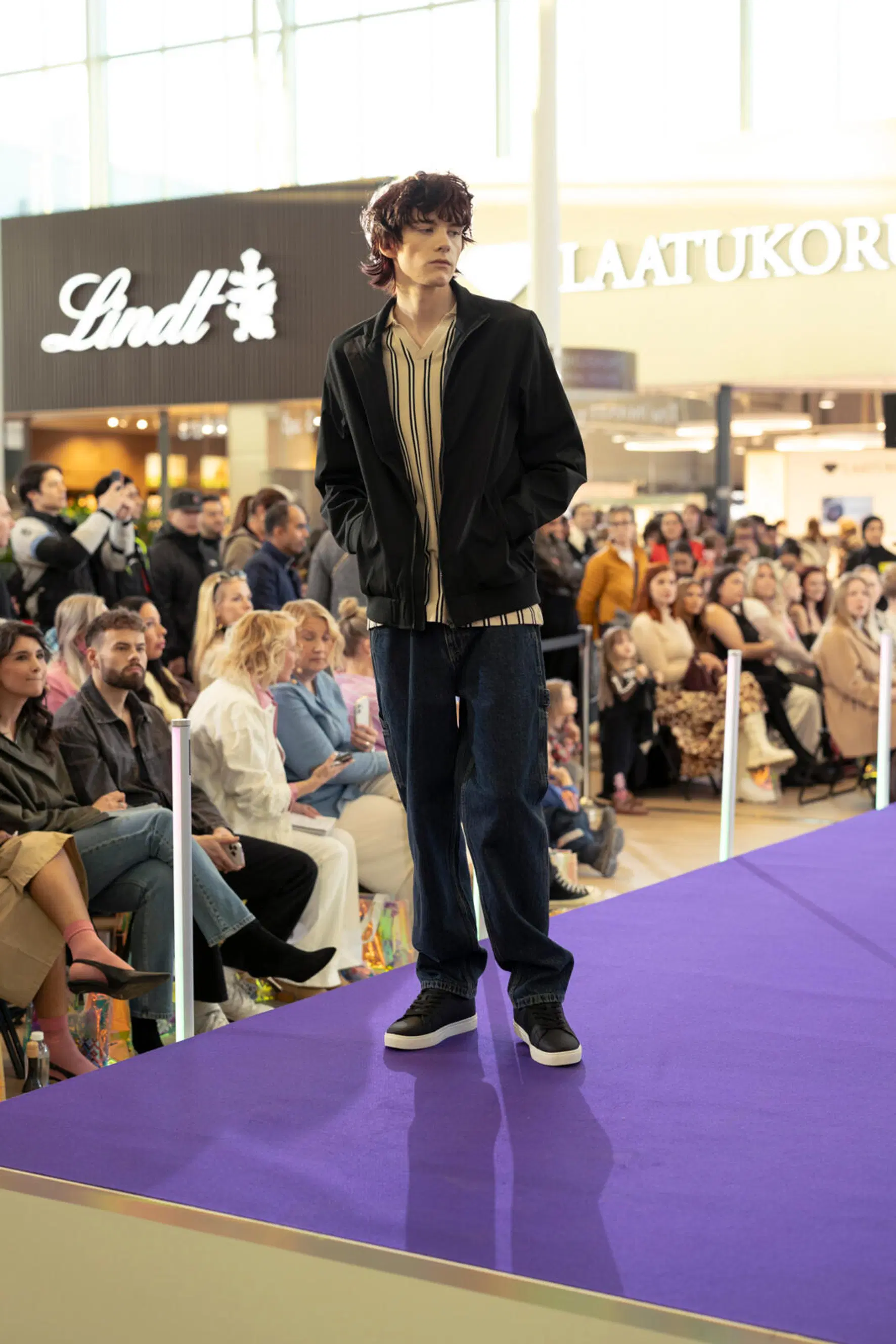 Miesmalli kävelee violetilla catwalkilla, musta takki, raidallinen paita ja tummansiniset farkut; yleisö taustalla.