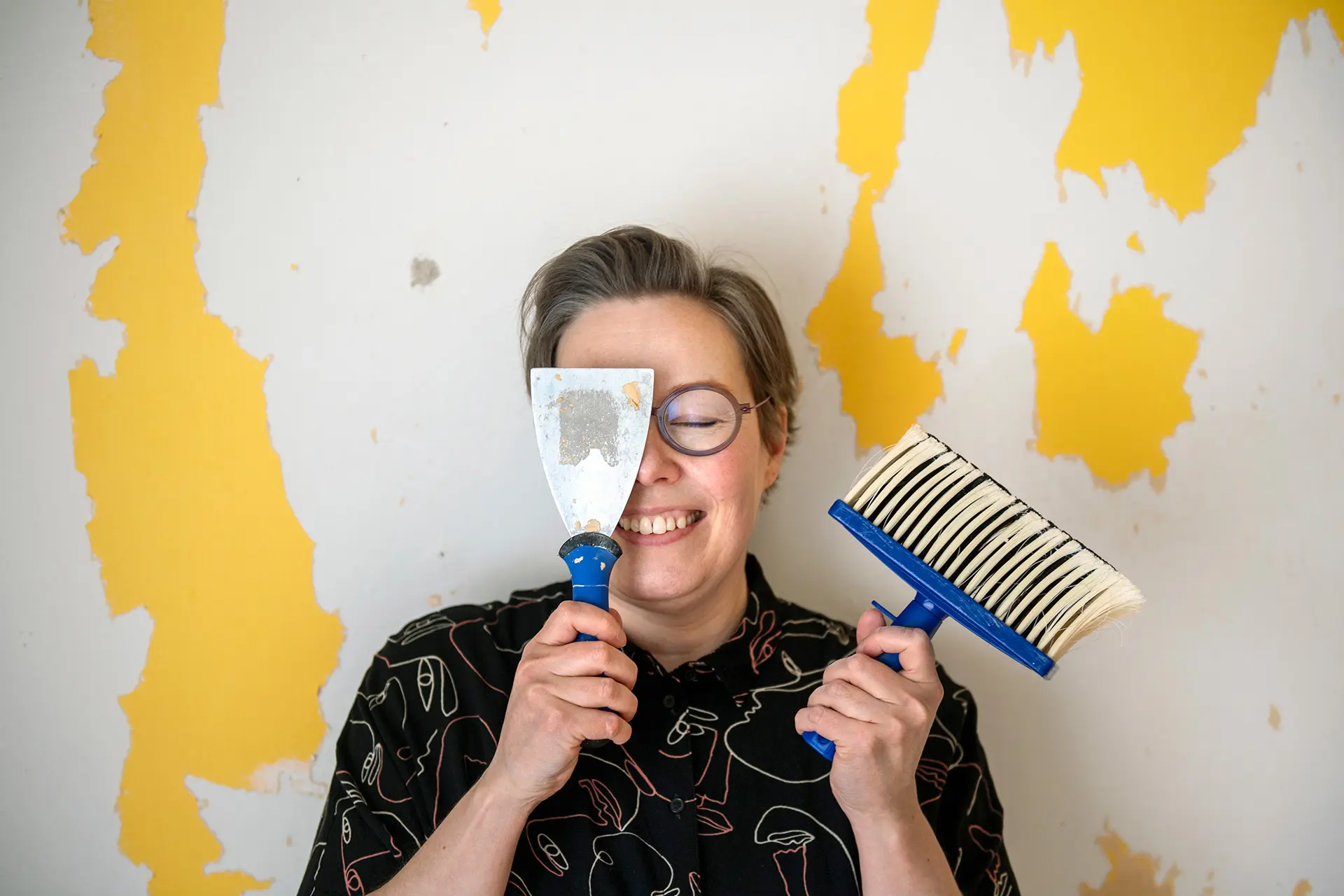 Kitti Suoranta silmät kiinni, kädessä tela ja pensseli, taustalla valkoista seinää, jossa keltaista maalia.