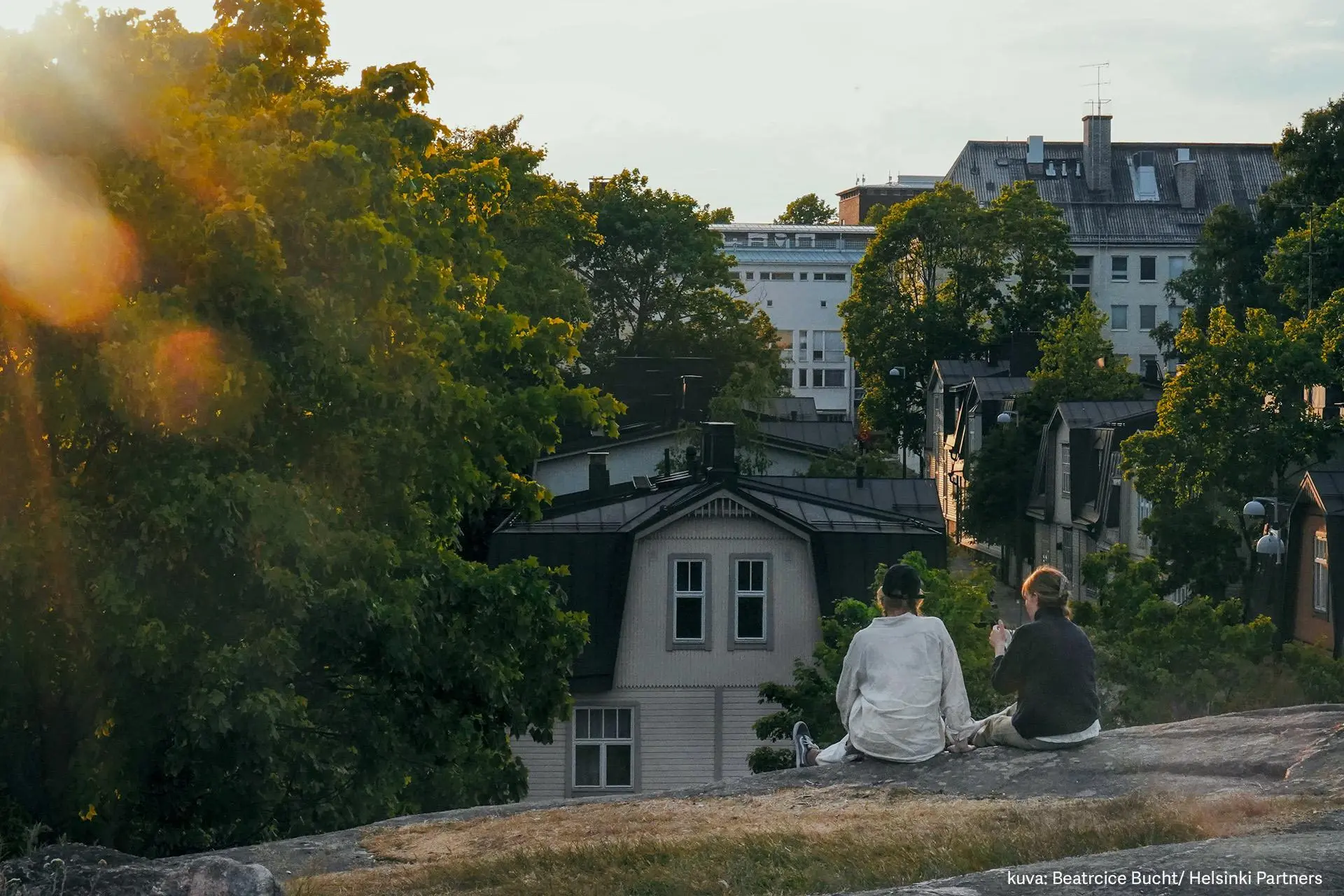 Kaksi ihmistä istuu selin kameraan Vallilan kallioilla ilta-auringossa.