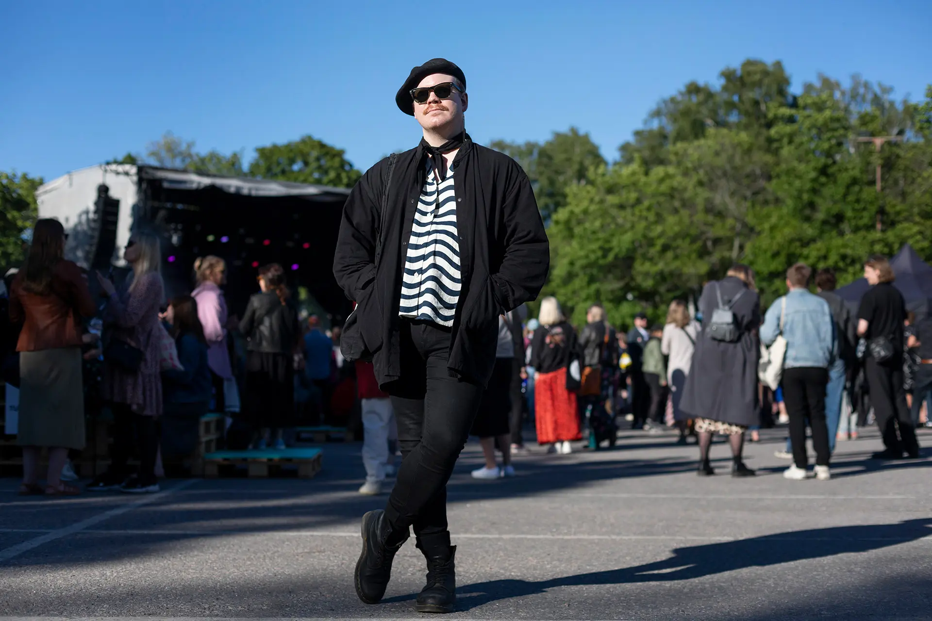 Antti Karjalainen seisoo tummassa asussa, aurinkolasit silmillä ja baskeri päässä, taustalla festarilava.