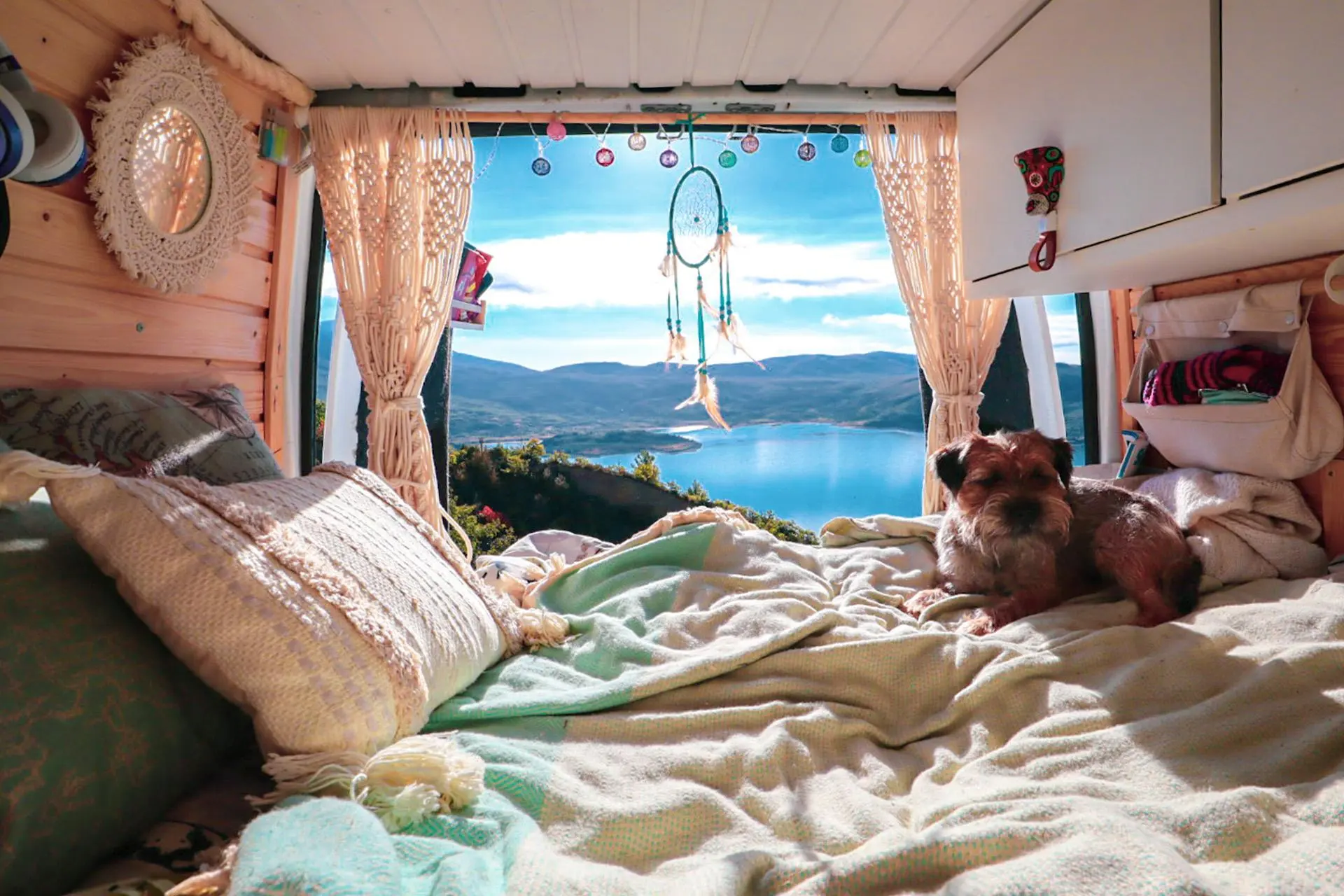 Kuva on otettu pakettiauton sisältä. Takaovet ovat auki. Etualalla on sänky, jolla koira makaa tyytyväisenä. Taustalla sininen taivas, meri ja vuoret.
