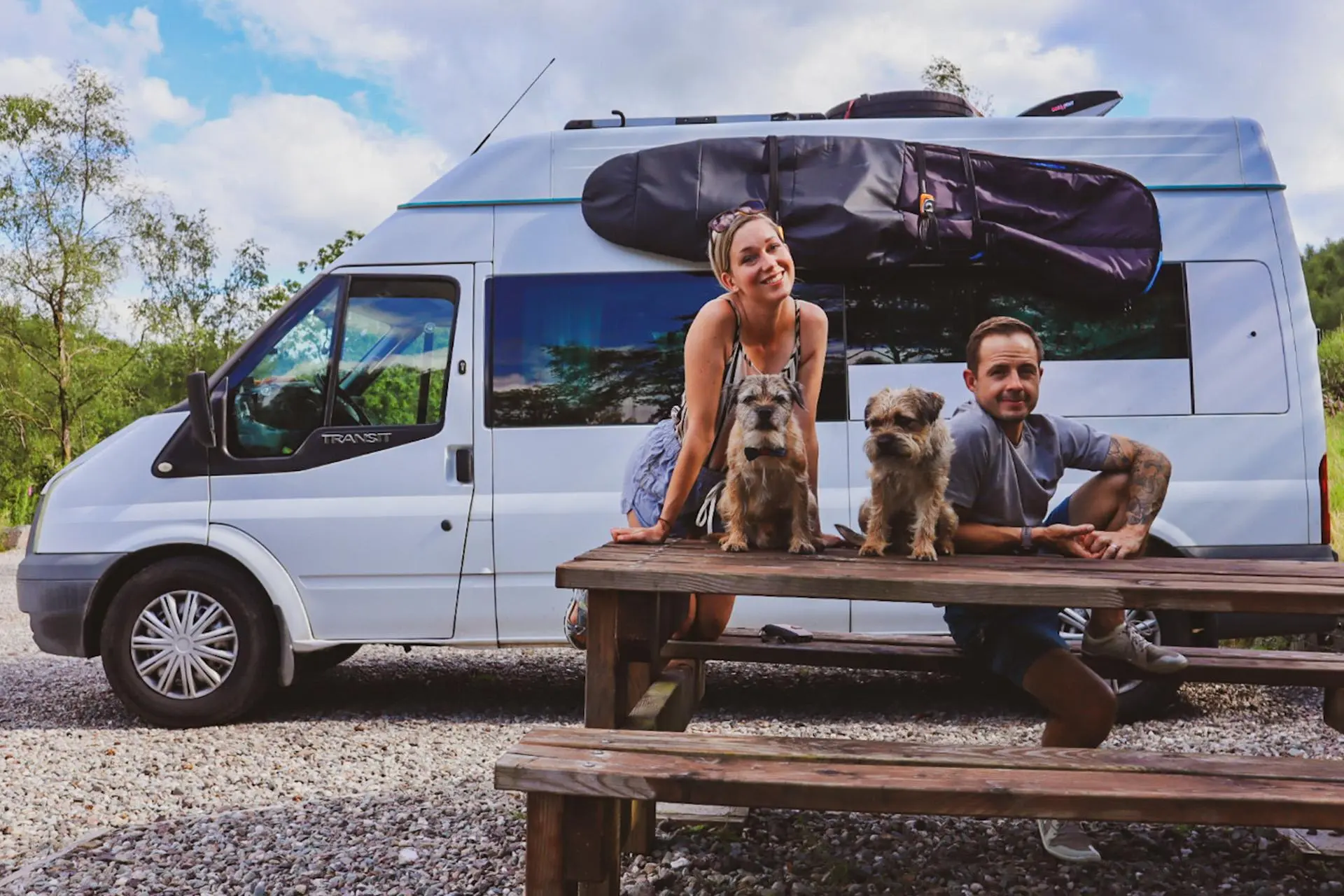 Etualalla puinen taukopaikan pöytä, jonka ääressä ovat Emma, Craig ja pariskunnan kaksi bordercollieta. Heidän takanaan on matkailuautoksi muuntautunut Ford Transit.