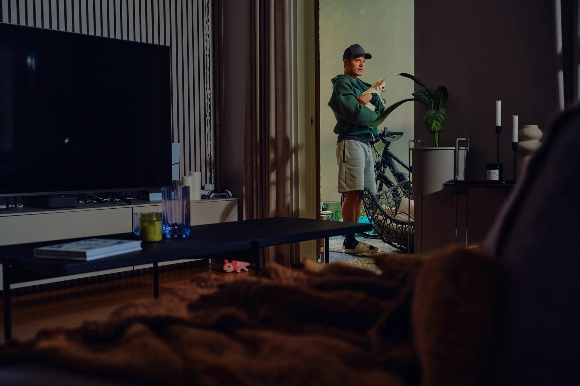 Niko Saarinen seivoo parvekkeella vaalea chihuahua sylissään. Parvekkeella näkyy myös korituoli ja polkupyörä. Etualalla näkyy olohuonetta, jossa on sohvapöytä, tv ja viherkasvi.