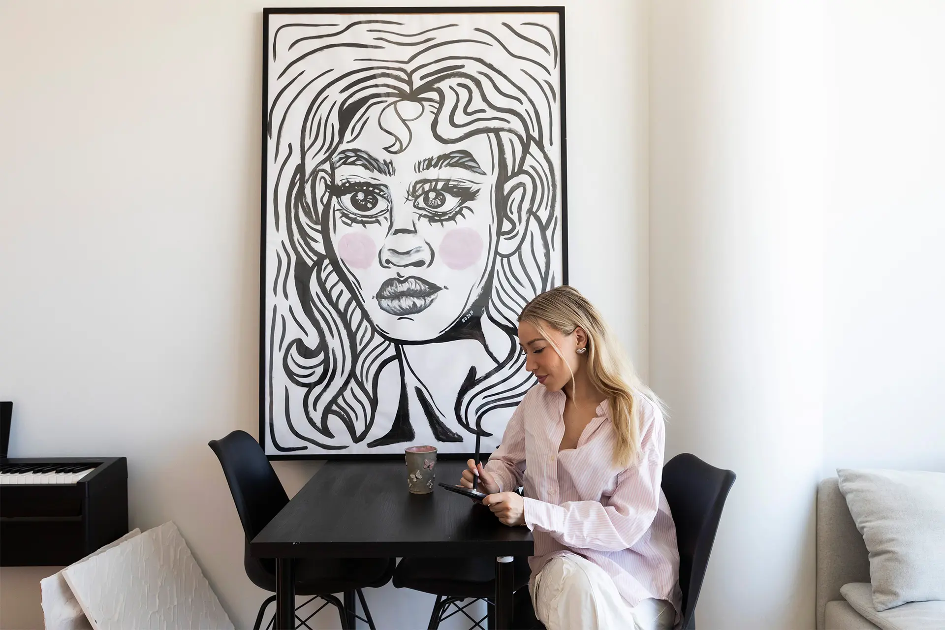 Nelli Orell istuu mustan pöydän ääressä ja piirtää, taustalla maalaus.