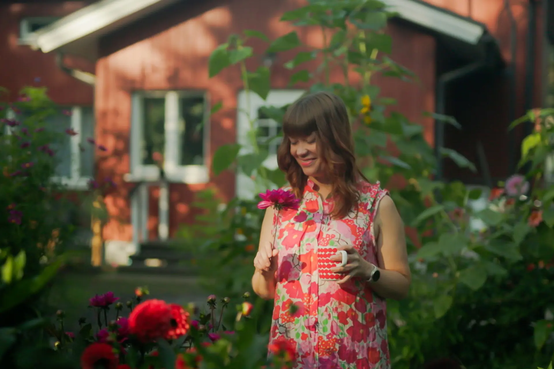 Reetta Mölsä puutarhassa kahvikupin kanssa, taustalla punainen talo.