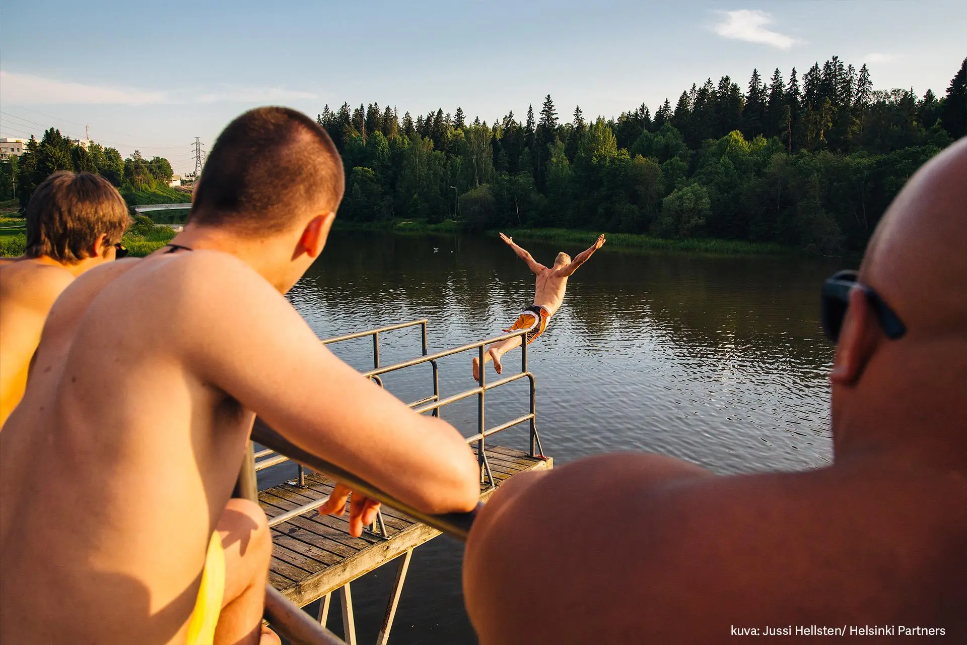 Nuoria miehiä selin kameraan katsomassa laiturilta veteen hyppäävää miestä.