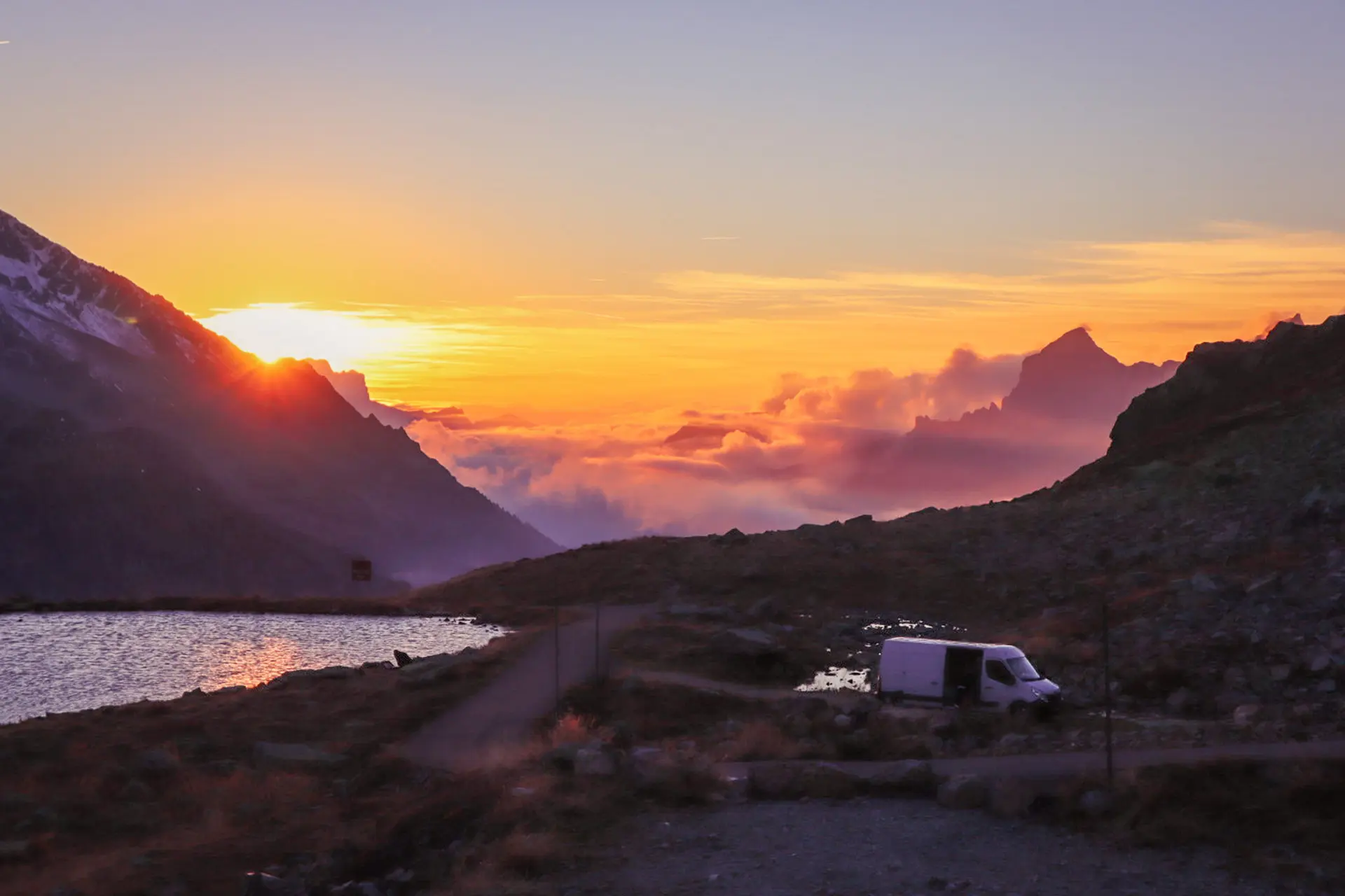 Näköala vuoristossa. Aurinko nousee vuorten takaa. Kuvassa etualalla pakettiauto ja pieni lampi.