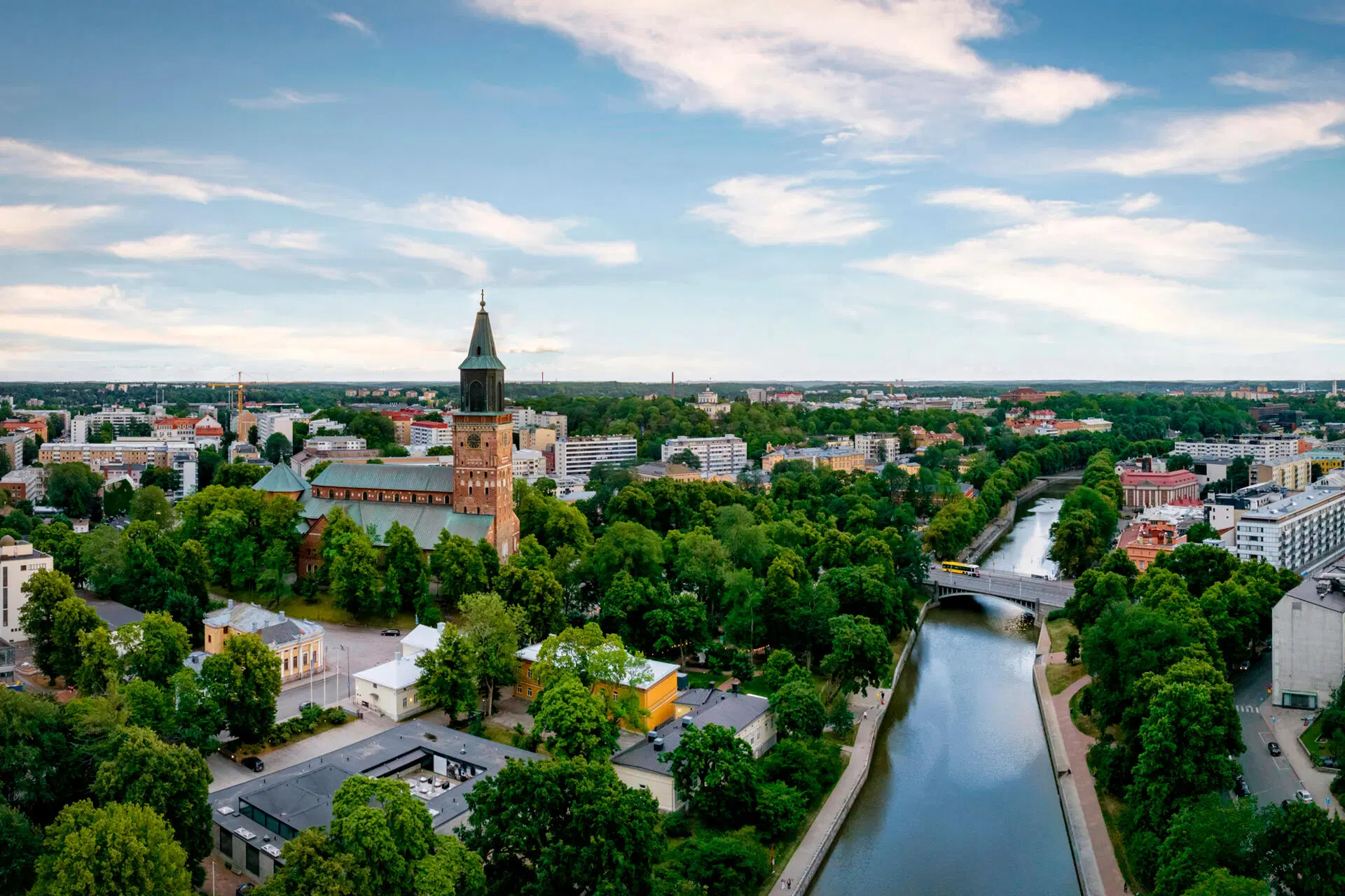 Ilmakuva Turusta, Aurajoki ja silta etualalla, vihreää puistoa, Turun tuomiokirkon torni kohoaa keskellä, sininen taivas.