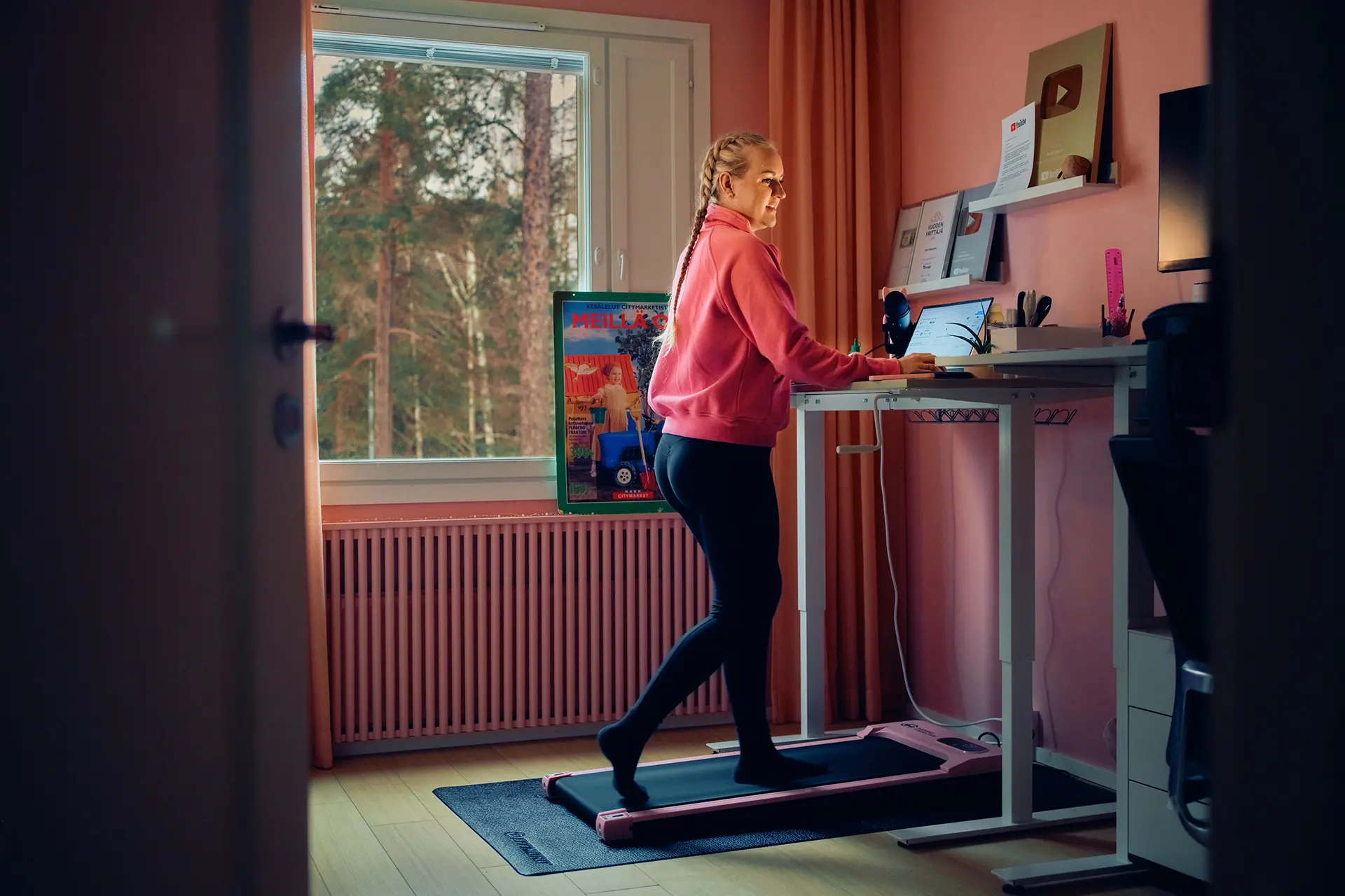 Auri Kananen seisoo kävelymatolla ja katsoo kannettavaa tietokonetta. Työhuone on maalattu pinkillä värillä.