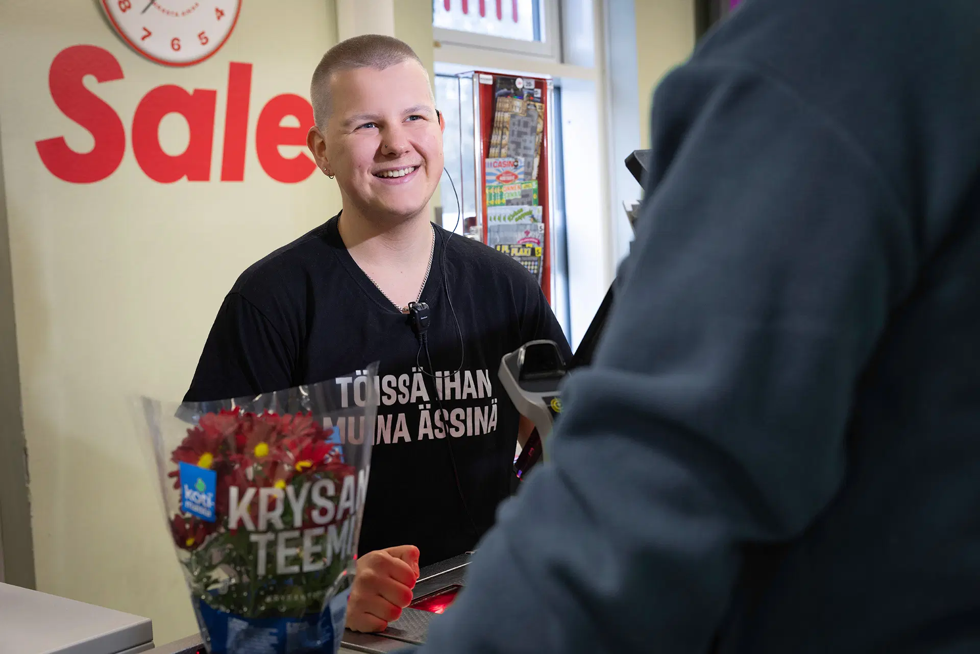 Kassatyöntekijä hymyilee ja skannaa ostoksia lähikaupassa; etualalla kukkakimppu.