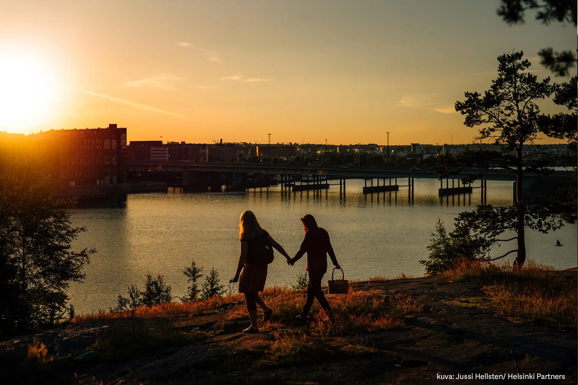 Kaksi henkilöä kulkee käsi kädessä piknik-korin kanssa Mustikkamaan kallioilla auringon laskiessa.