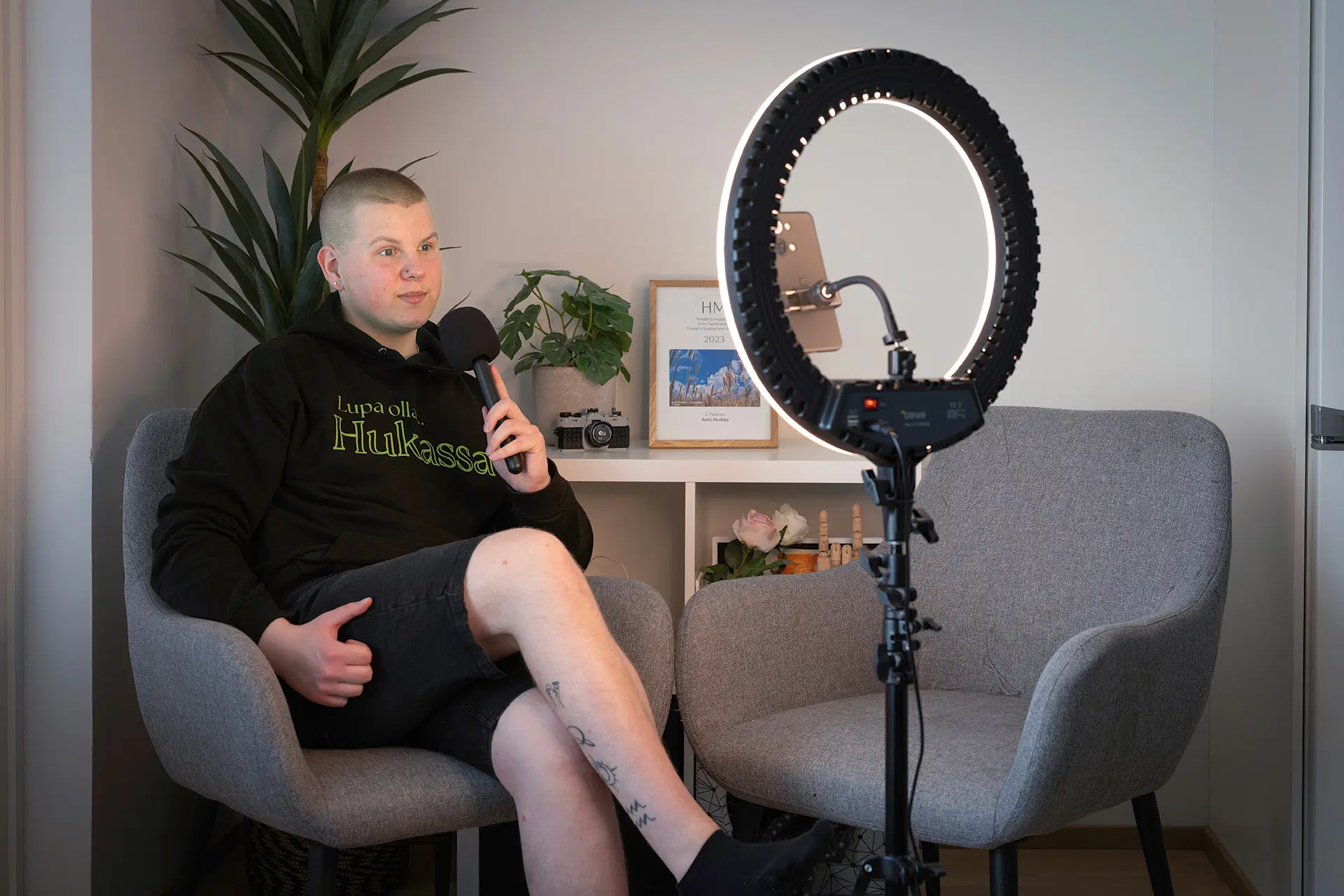 Nuori henkilö nauhoittaa podcastia mikrofonin kanssa, rengasvalo ja kamera edessä, vaalea huone ja kaksi harmaata tuolia.