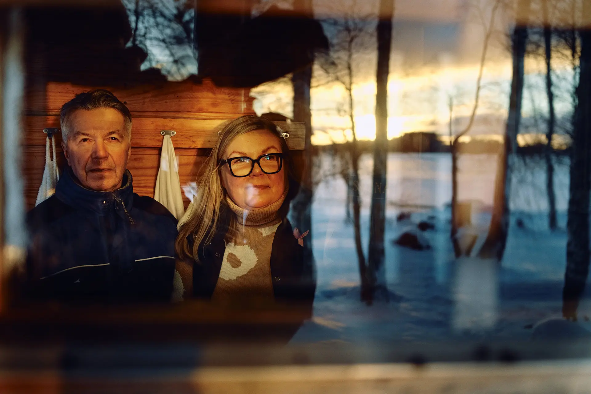 Olli ja Liisa Kalliokoski ikkunan läpi kuvattuna. Ikkunan heijastuu myös luminen maisema.
