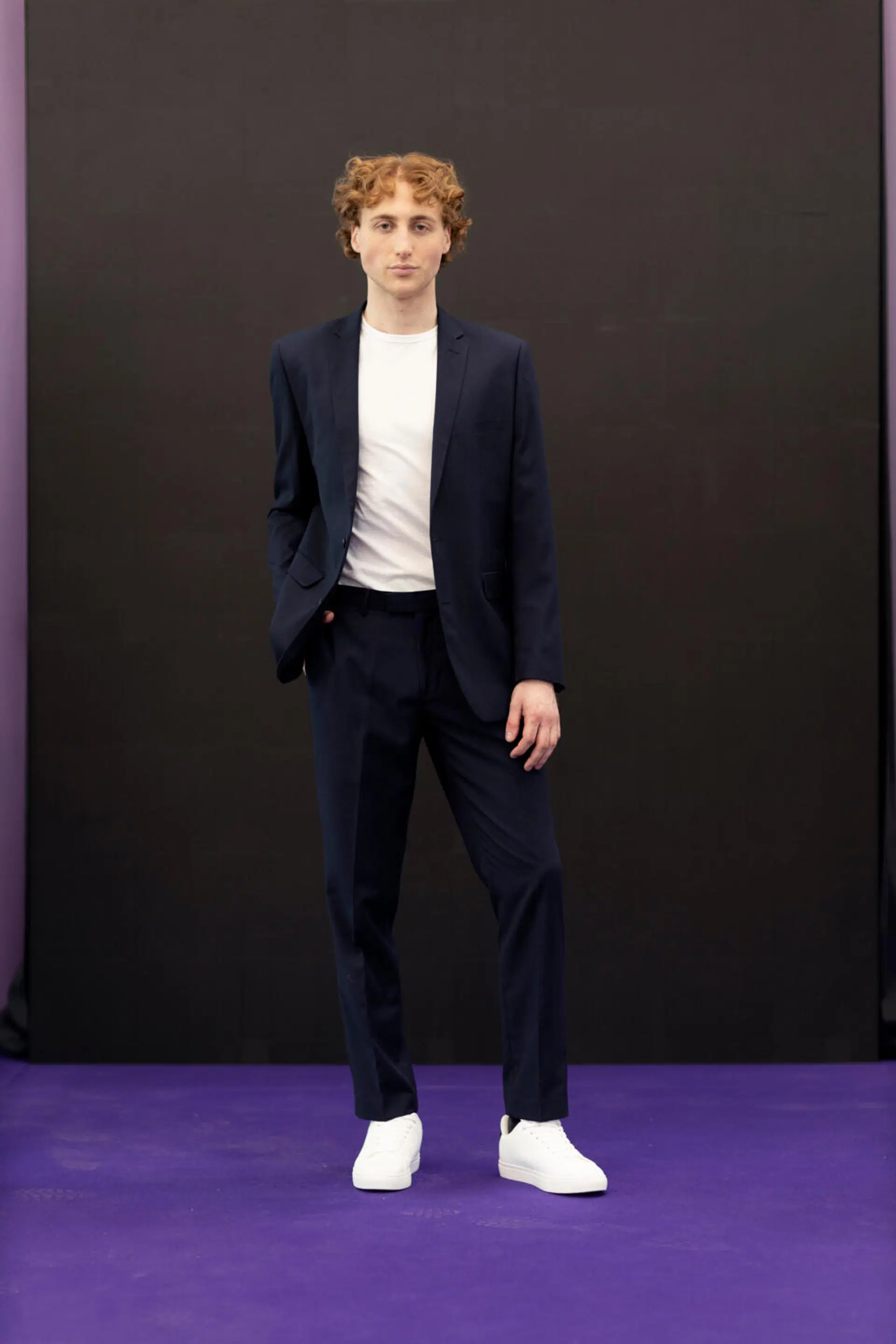 Henkilö seisoo violetilla lattialla tummassa puvussa, valkoinen t-paita ja valkoiset lenkkarit, musta tausta.