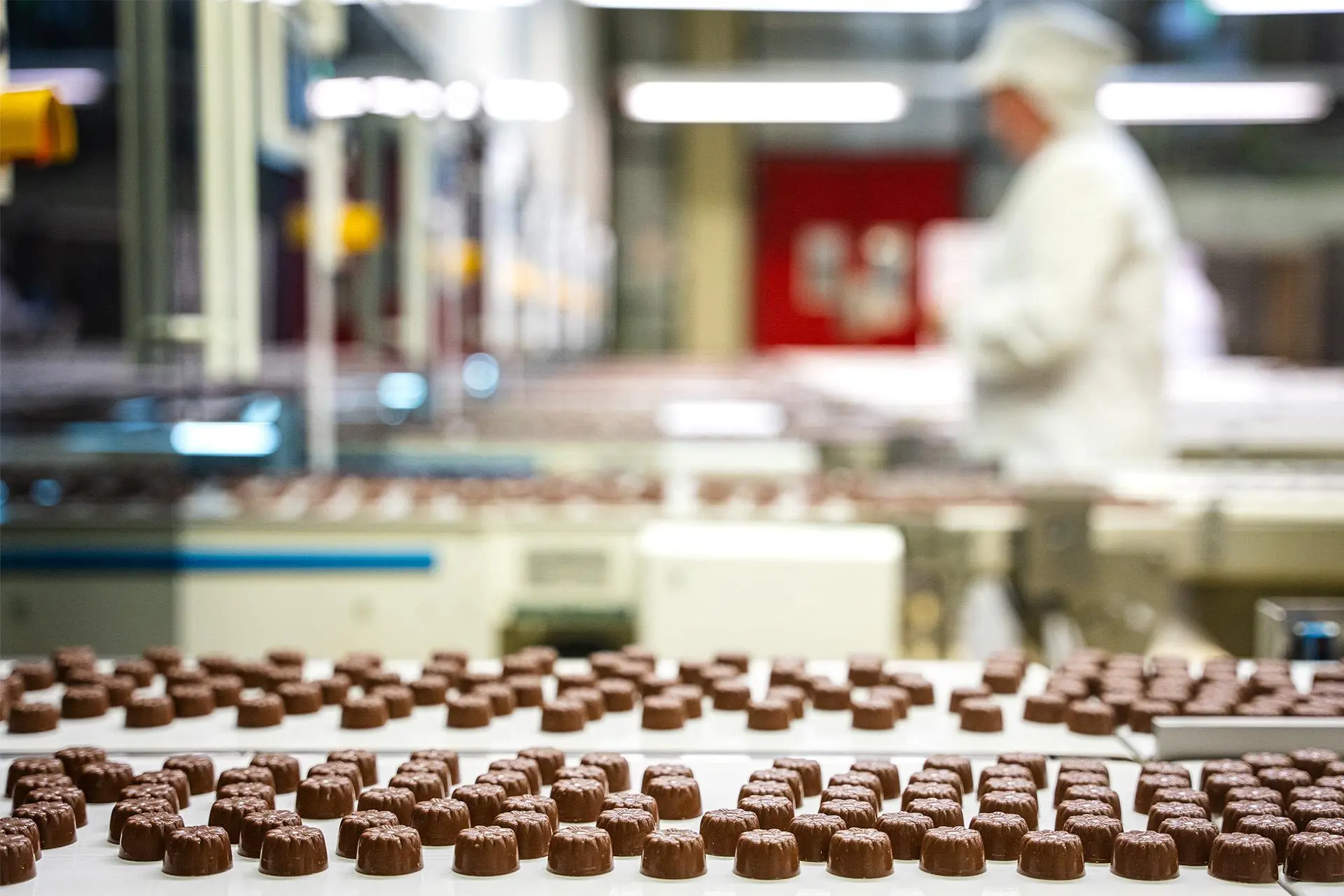 Yksittäisiä suklaakonvehteja tehtaan hihnalla. Taustalla kävelee työntekijä.