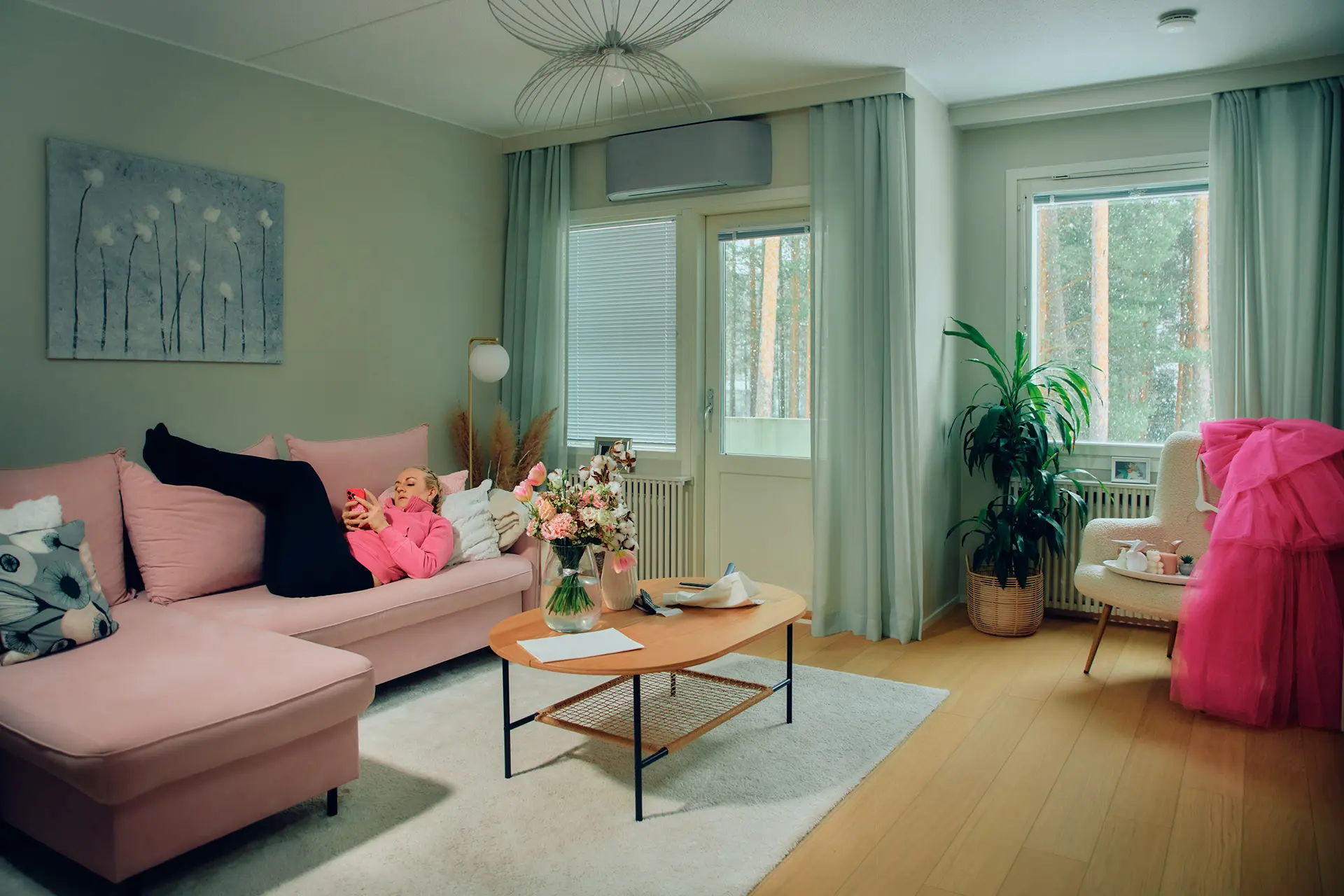 Auri Kananen selaa puhelinta pinkillä sohvalla maaten. Olohuoneessa on myös puinen sohvapöytä, jolla on paljon kukkia, suuri kukka-aiheinen taulu ja ikkunat, joista näkyy metsämaisema.