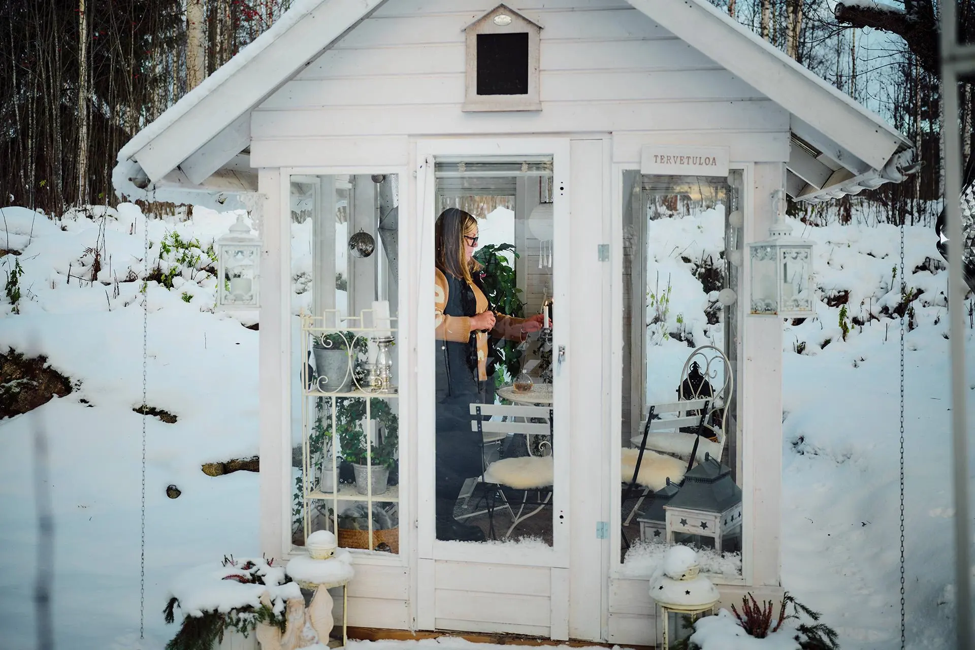 Liisa Kalliokoski pienessä piharakennuksessa, jossa on kasveja, pöytä ja muutama tuoli.