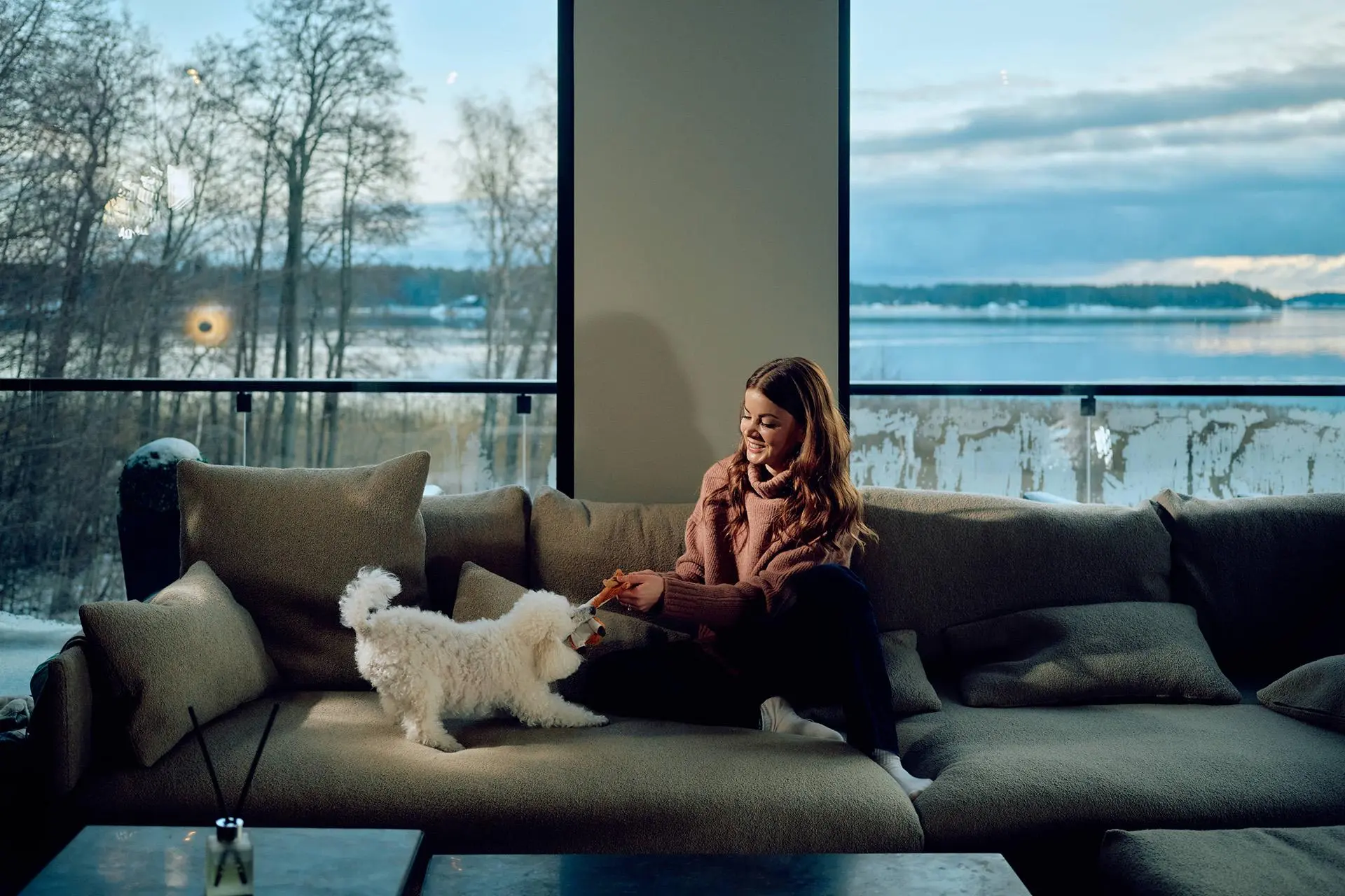 Janni Hussi ja pieni koira leikkivät sohvalla, jonka takana olevista suurista ikkunoista näkyy merelle.