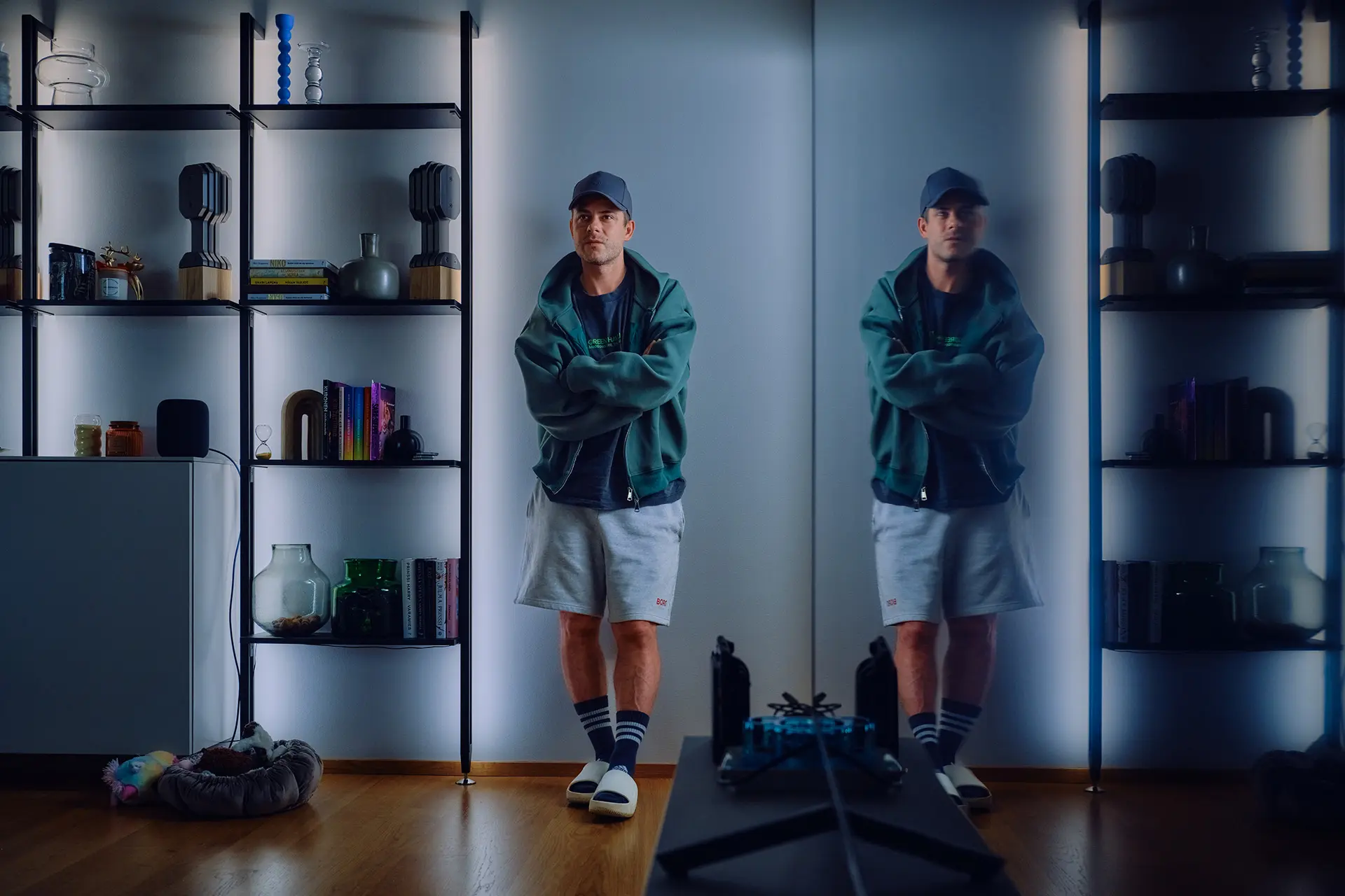 Niko Saarinen nojaa seinään. Vieressä on hyllykkö, jossa on palkintoja, kirjoja ja koriste-esineitä. Etualalla oikealla näkyy pimeä tv-ruutu, josta huone heijastuu.