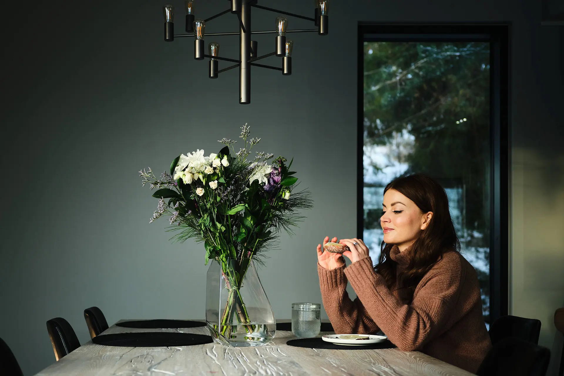 Janni Hussi syö leipää pöydän ääressä. Pöydällä on kukkakimppu ja katossa moderni valaisin.