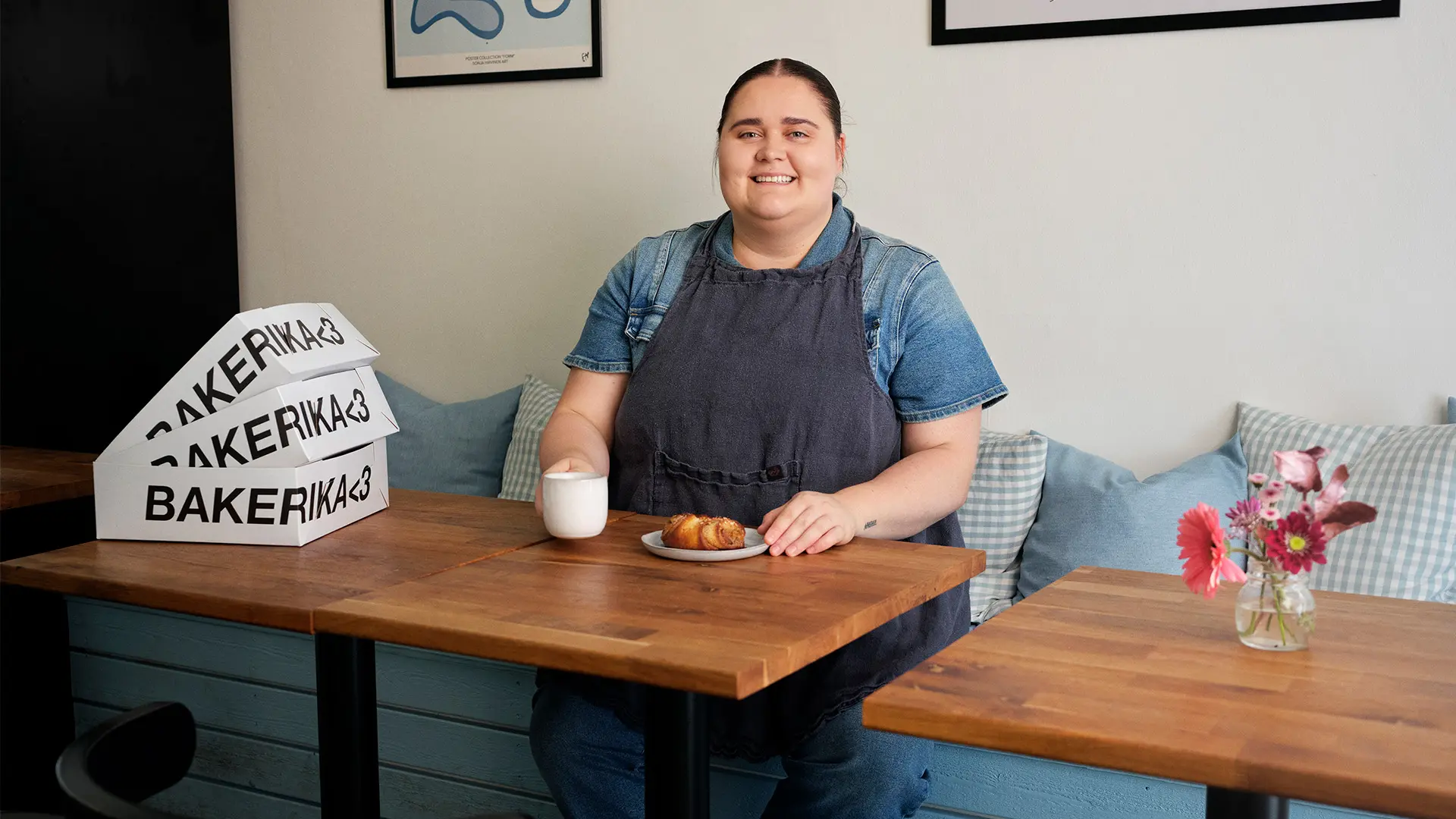 Erika Poussa istuu kahvilapöydän ääressä. Pöydällä on kahvikuppi, korvapuusti, kukkamaljakko ja leivoslaatikoita.