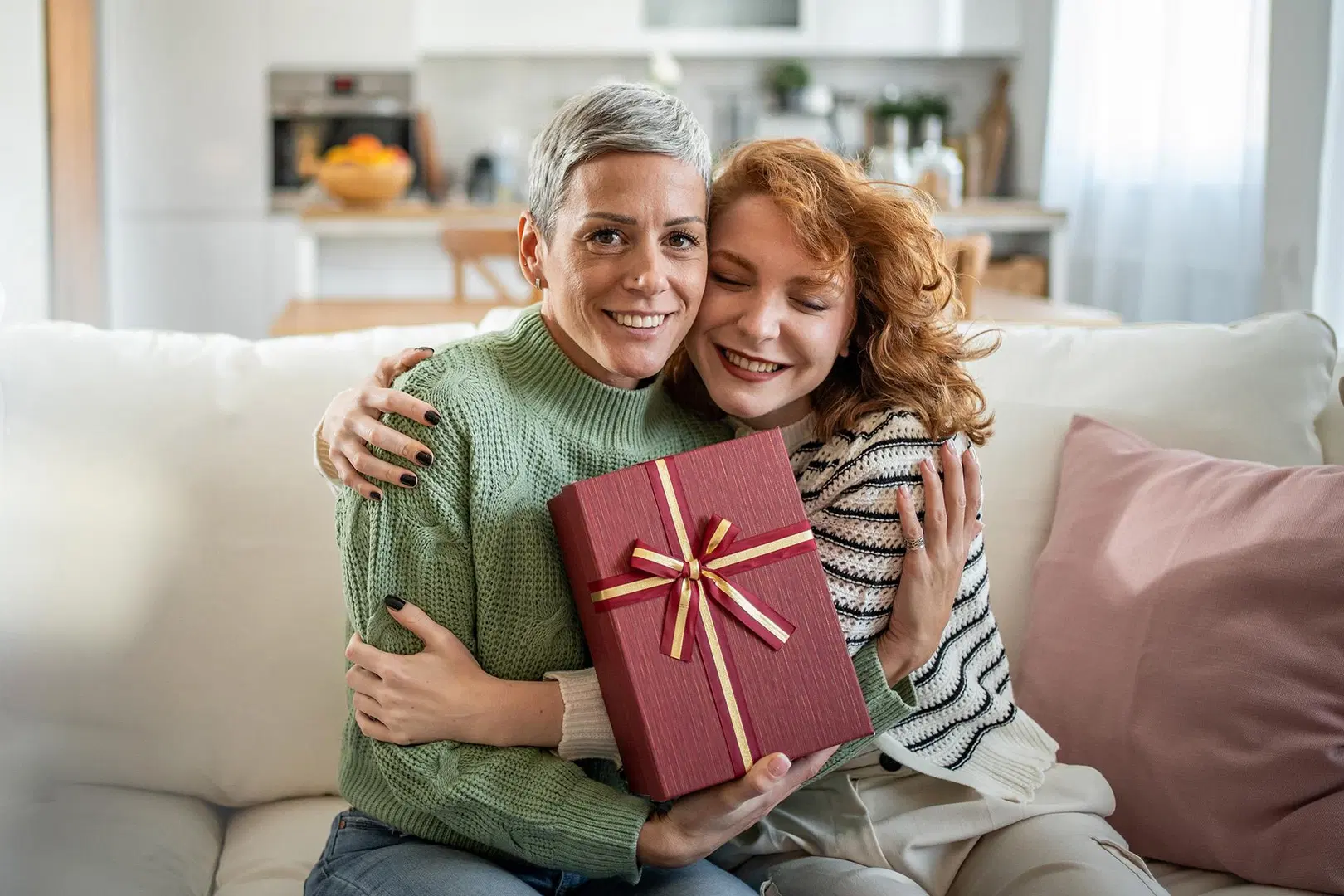 Harmaahiuksinen nainen antaa paketin nuoremmalle punahiuksiselle naiselle ja nuorempi nainen halaa. 