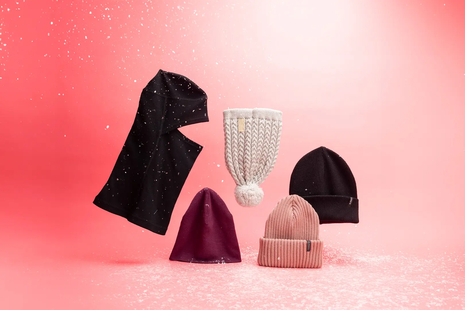 Lumisadetta ja viisi erilaista pipoa vaaleanpunaisella taustalla. Osa pipoista leijuu ilmassa.