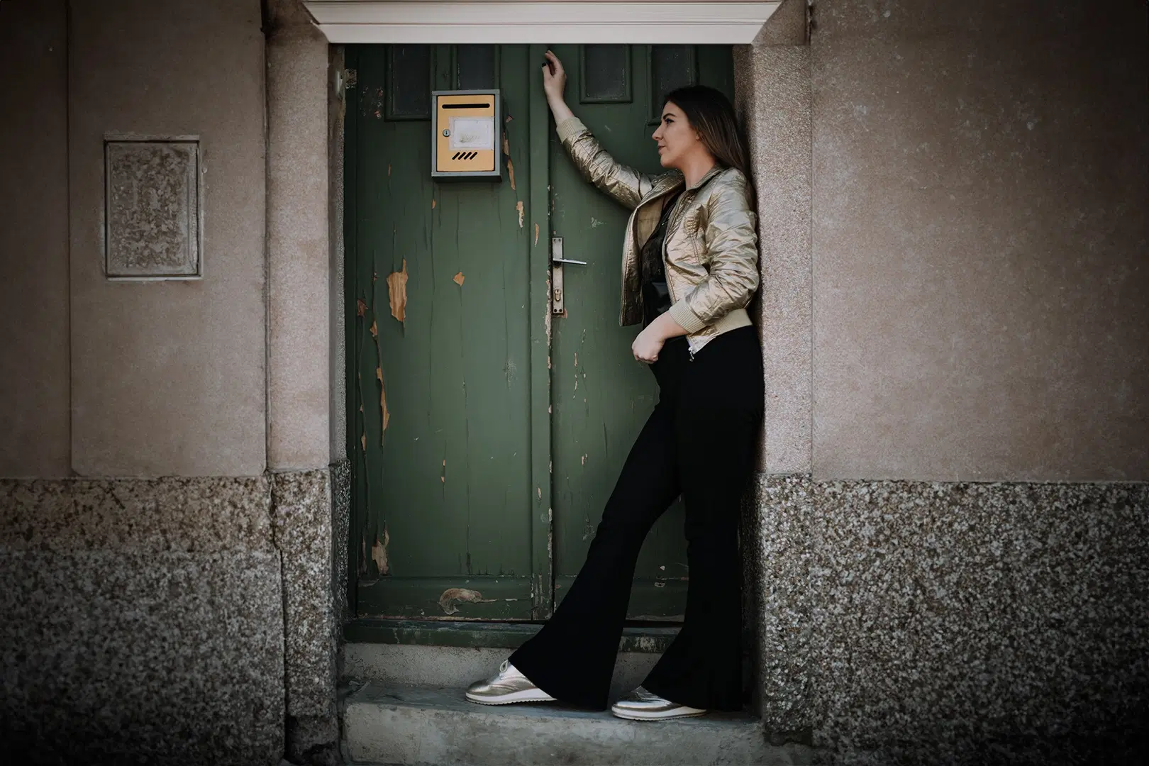 Nainen seisoo vihreän oven edessä, jalassa mustat trumpettimalliset housut.
