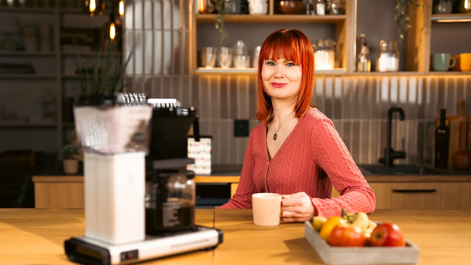 Punahiuksinen nainen istuu kahvikuppi kädessään. Pöydällä hänen edessään on kahvinkeitin ja hedelmiä vadissa.