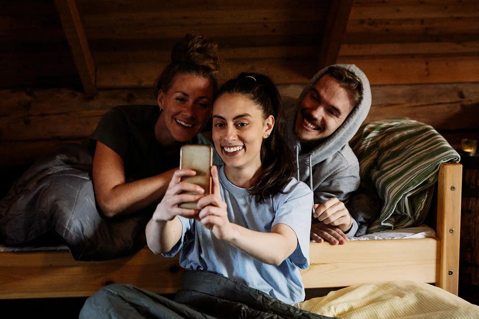 Kolme ystävää sängyllä mökissä, hymyilevät ja ottavat selfien puhelimella.