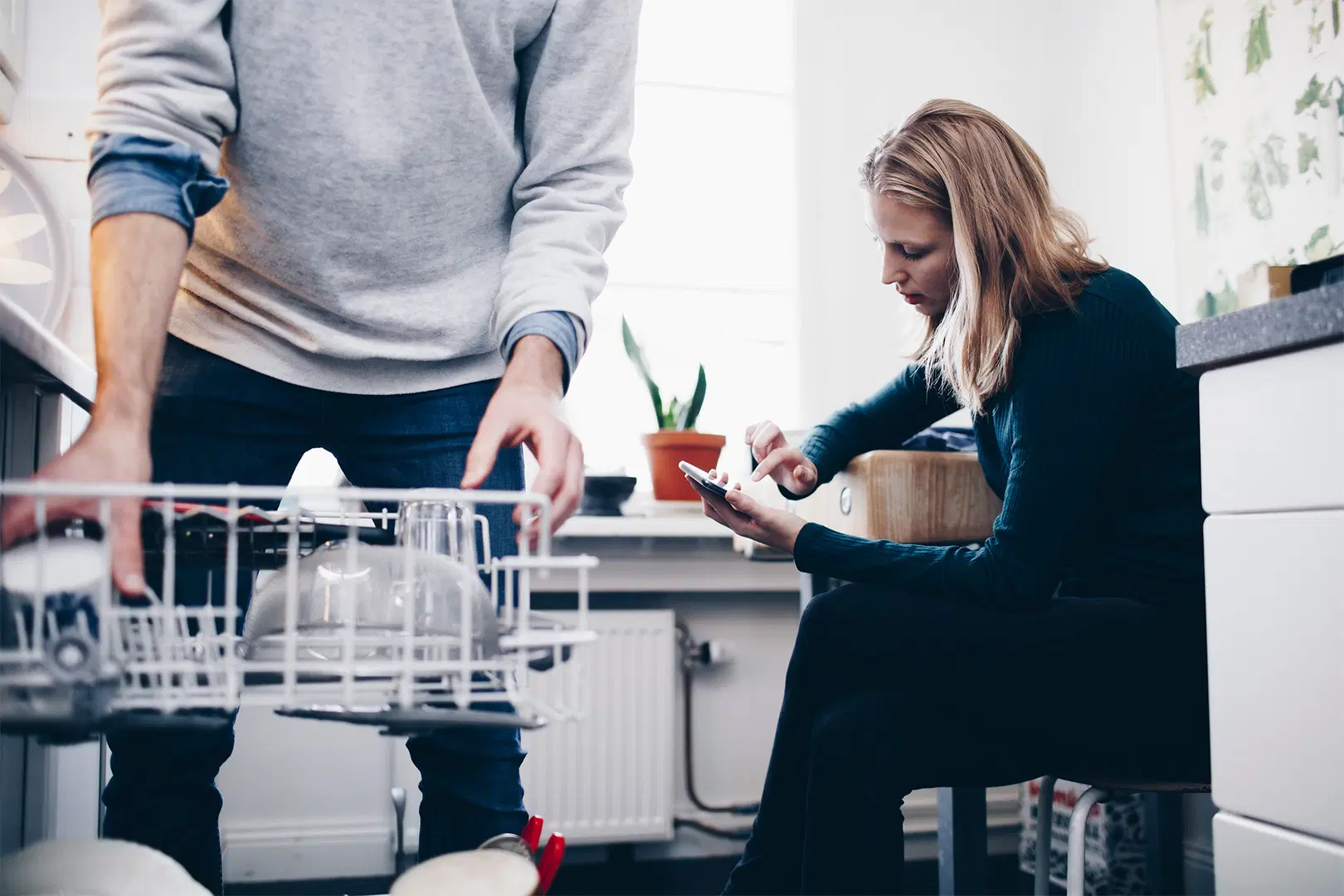 Mies täyttää astianpesukonetta ja nainen istuu taustalla selaillen puhelintaan.