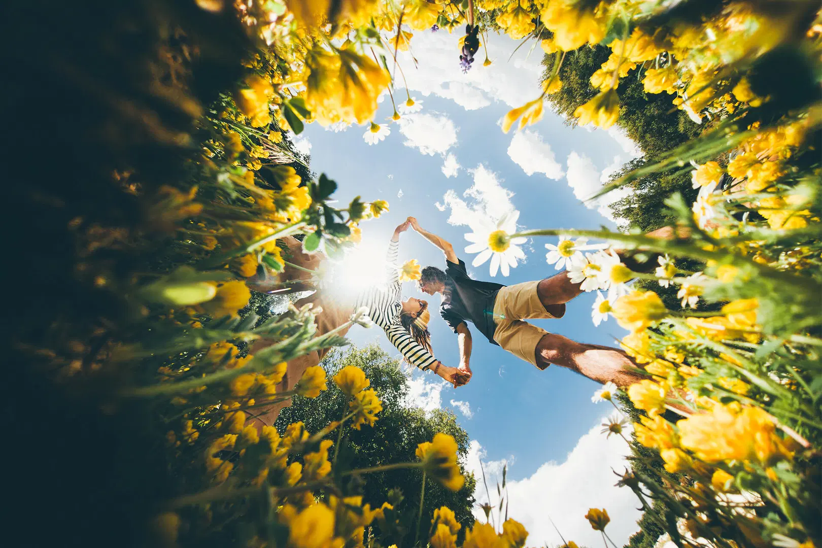 Kaksi ihmistä hyppii keltaisten kukkien keskellä, aurinko paistaa ja taivas on sininen pilvineen.