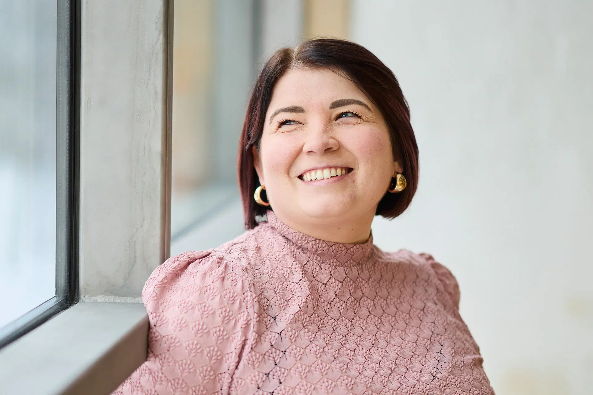 Sanna Latvala kuvattuna ikkunan äärellä, katsoo ikkunasta ulos ja hymyilee.